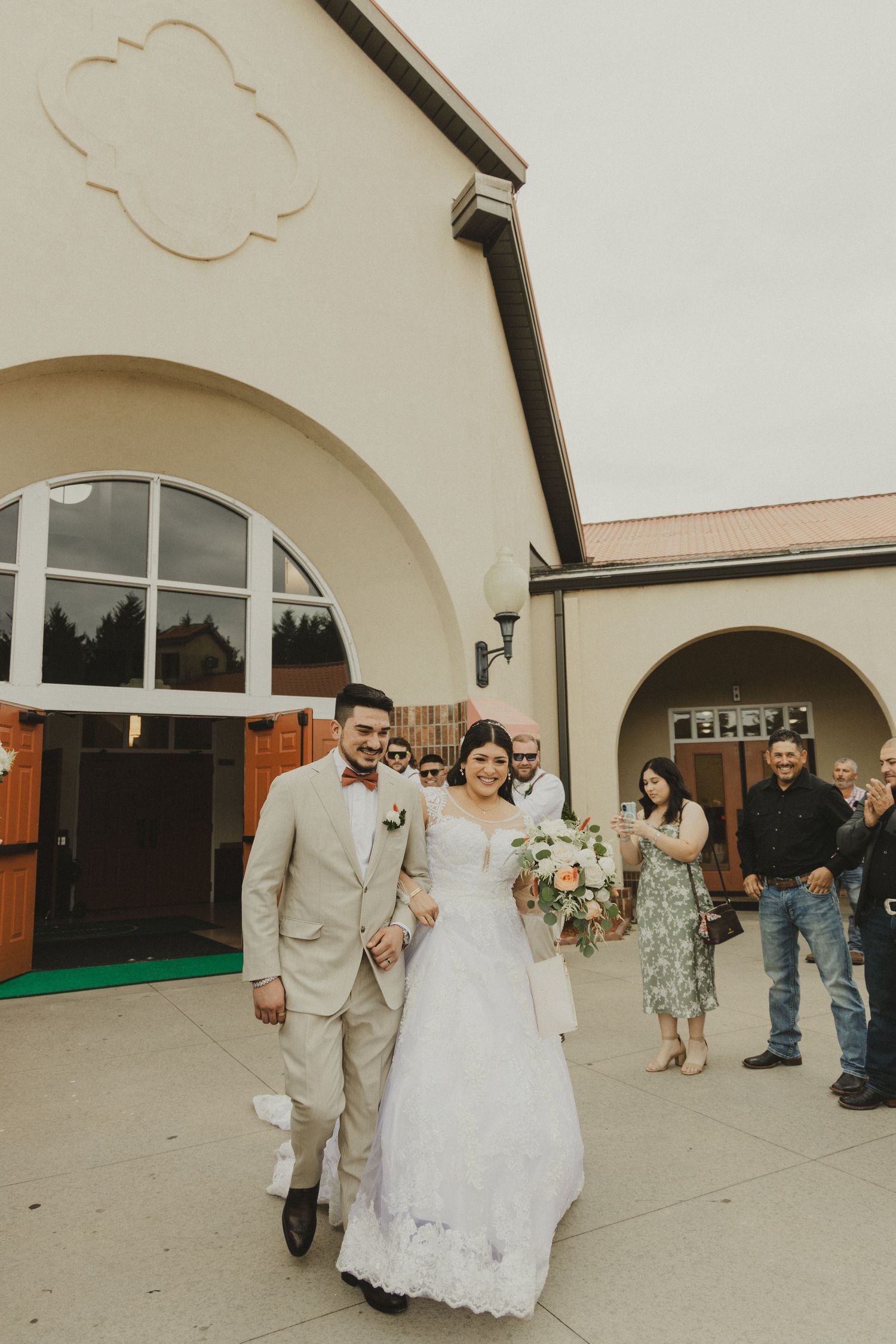 Bride and groom make their joyful exit from a beige arched church building with wedding guests looking on at their special day.