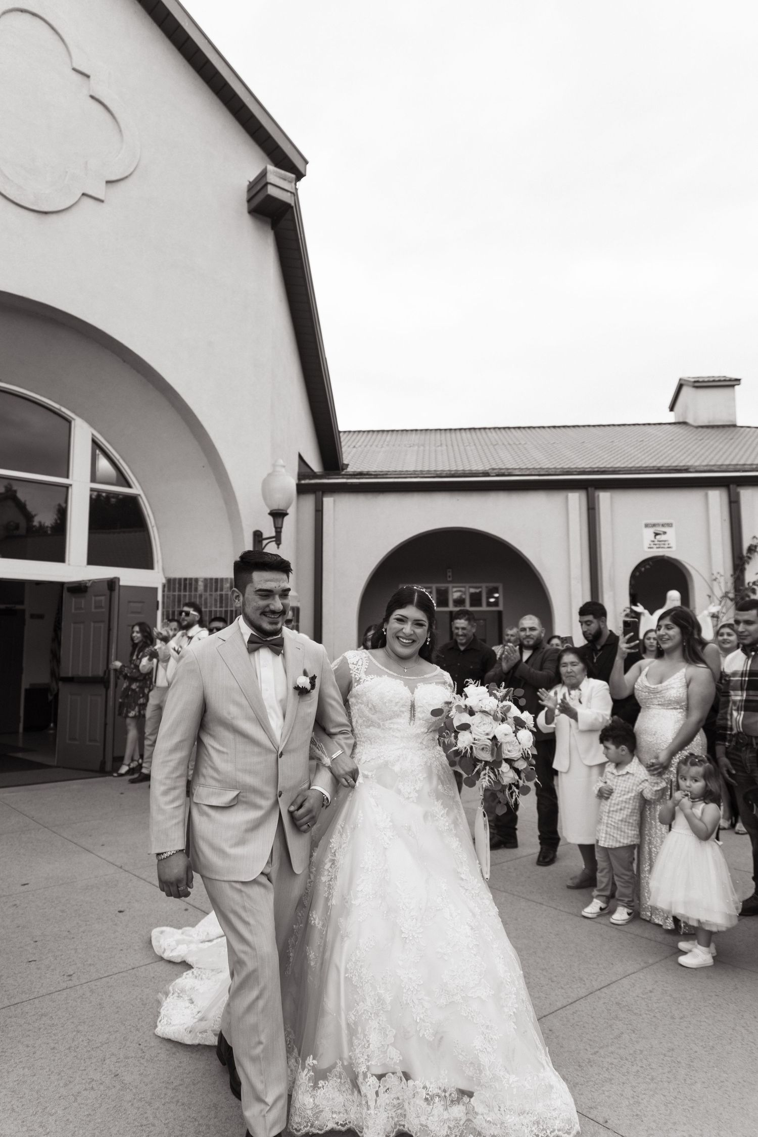 New married couple exits building surrounded by wedding guests in black and white wedding photograph.