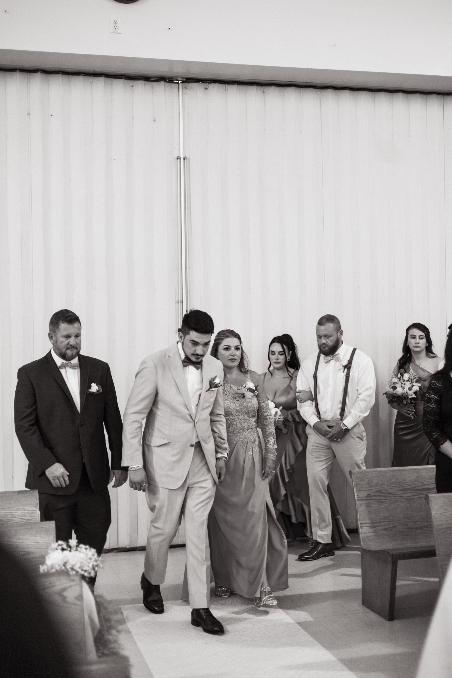 A wedding party walks together in a rustic venue with white walls, featuring a mix of formal attire and suspenders.