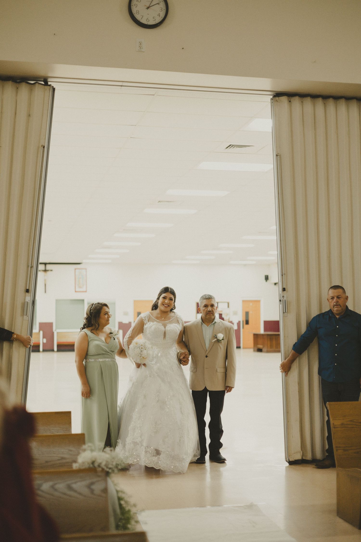 A bride in white gown stands with wedding party members in a bright hallway framed by beige curtains.