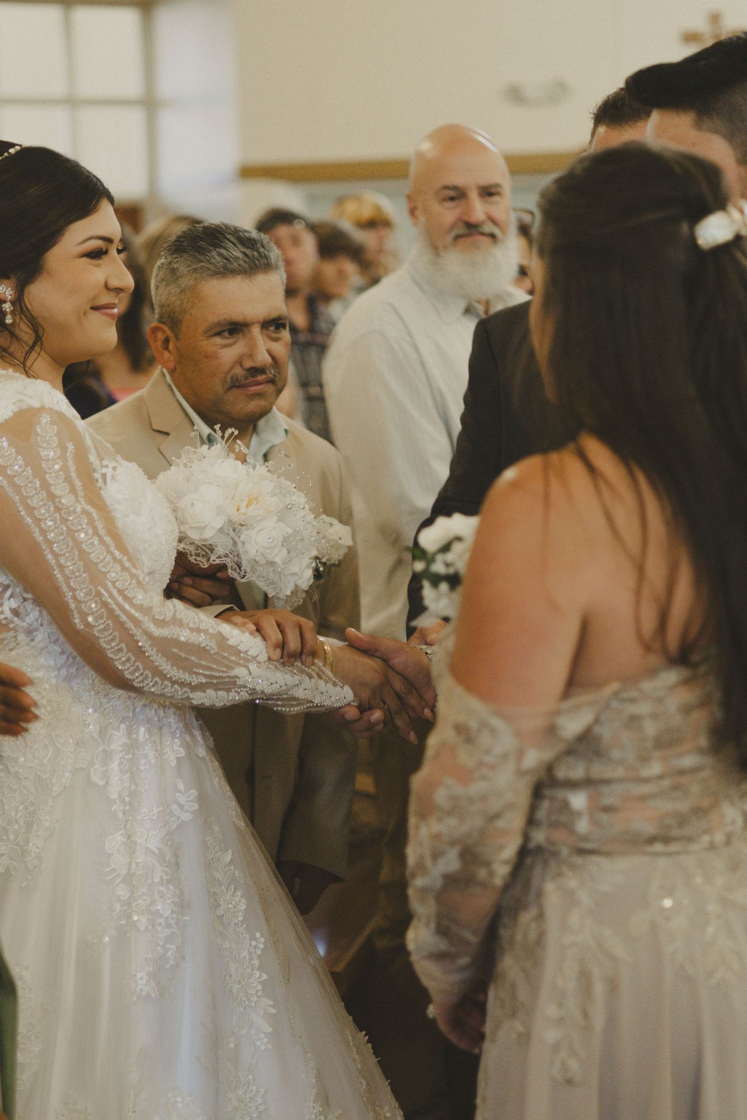 A bride in a white lace dress greets guests during a wedding reception while others look on in a warm indoor setting.