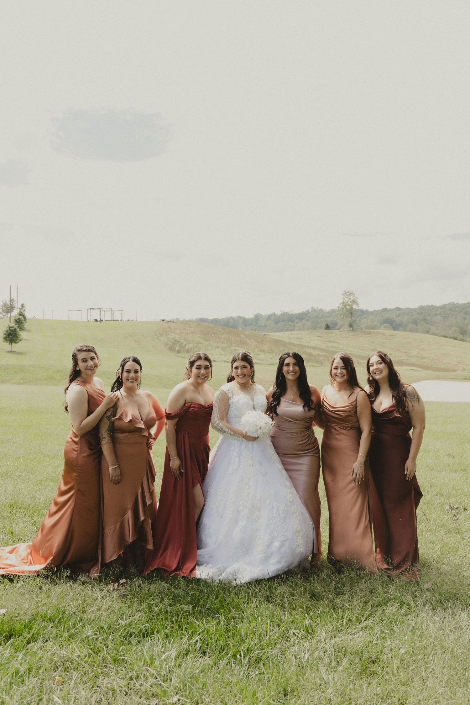 Bridal party wearing earthy brown tones poses together on grassy hillside for elegant wedding photos.
