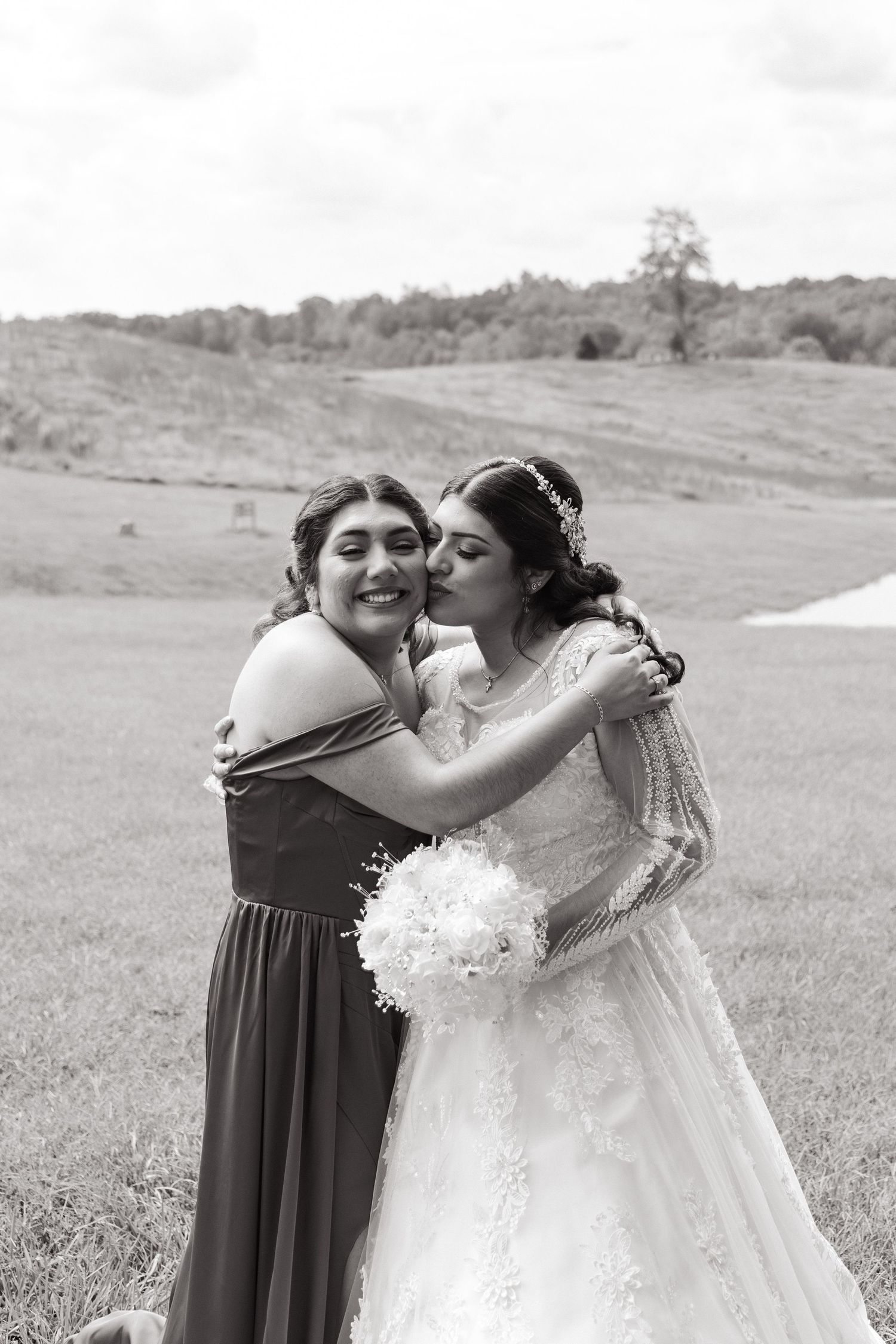 Black and white wedding photo showing two women embracing outdoors, one in a wedding dress holding a bouquet.