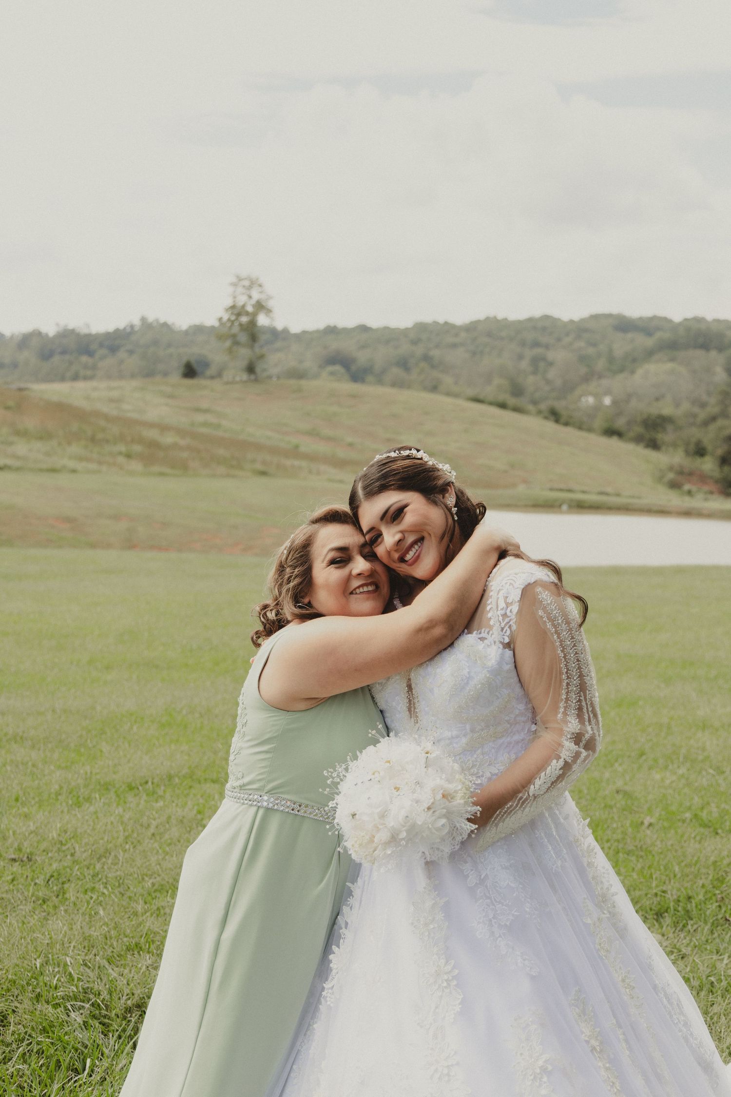 Two wedding attendees embrace on a grassy hillside with rolling hills and a lake in the background.