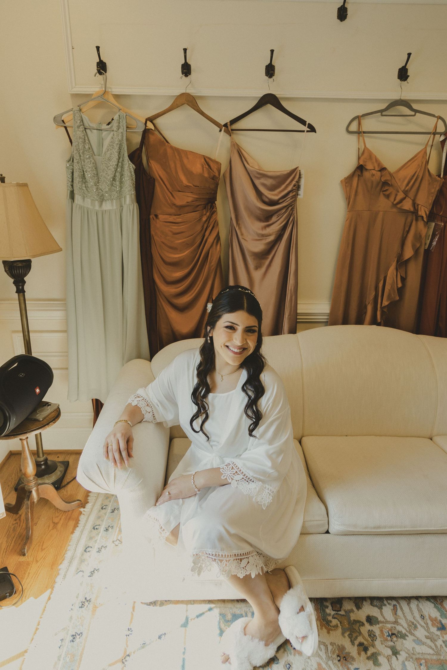 A collection of earth-toned bridesmaid dresses hanging on a clothing rack beside white robes on a cream couch.