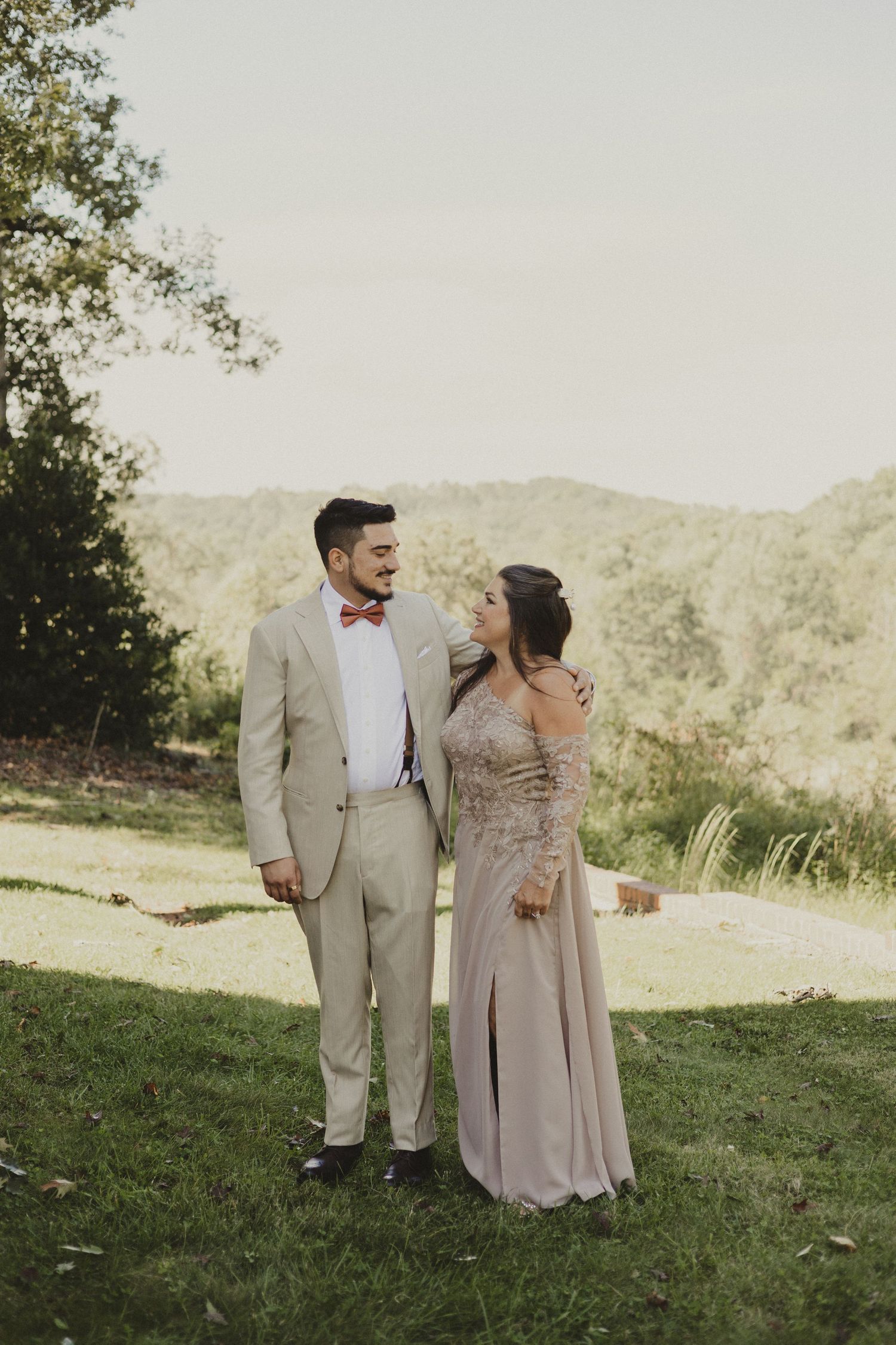 A couple poses together on a scenic hillside wearing formal attire at an outdoor event.
