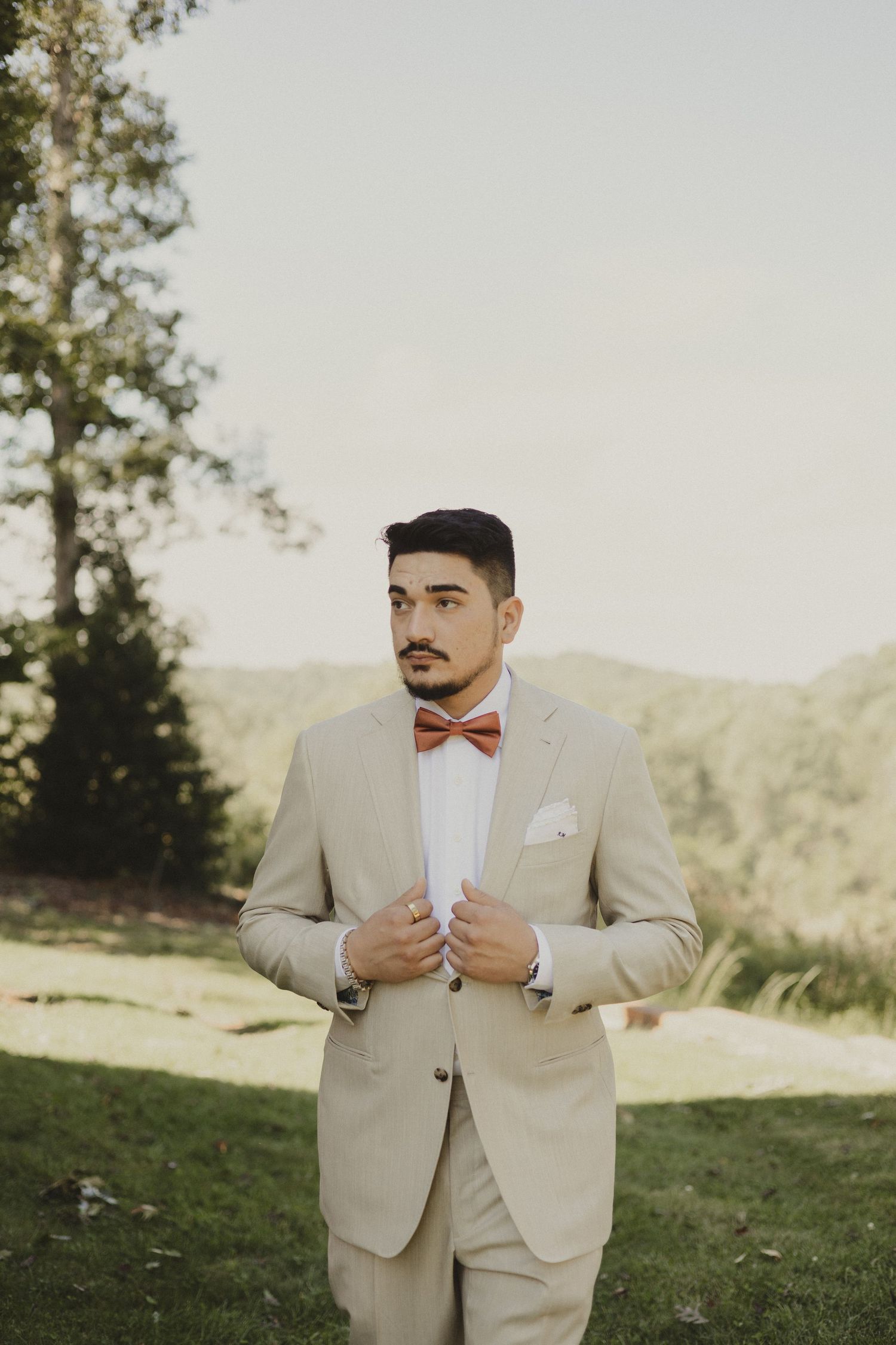 A person in a beige suit adjusts their tie while standing on a grassy hillside in natural sunlight.