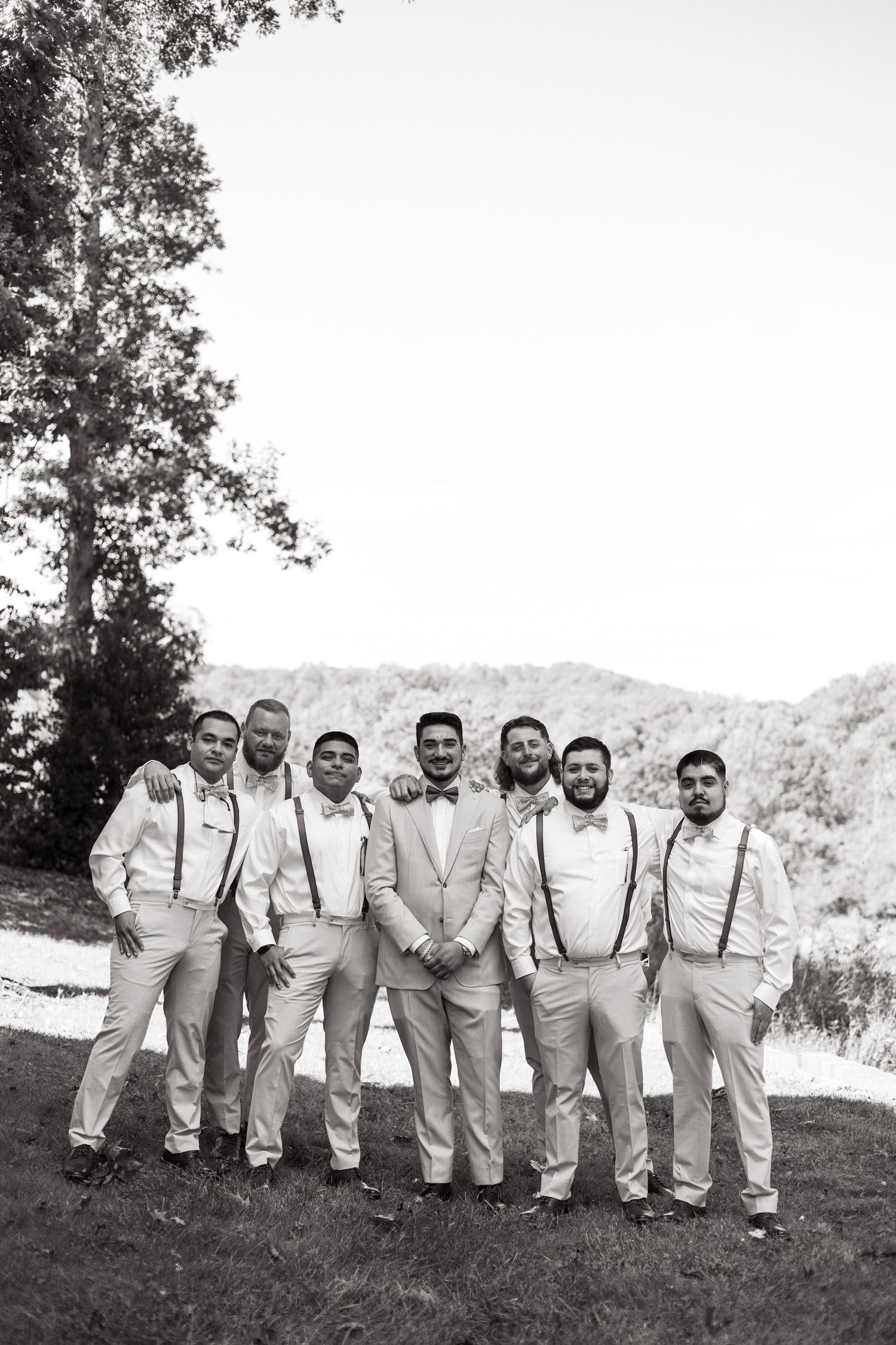 A black and white photo of groomsmen in light colored suits and suspenders posing outdoors on a hillside.