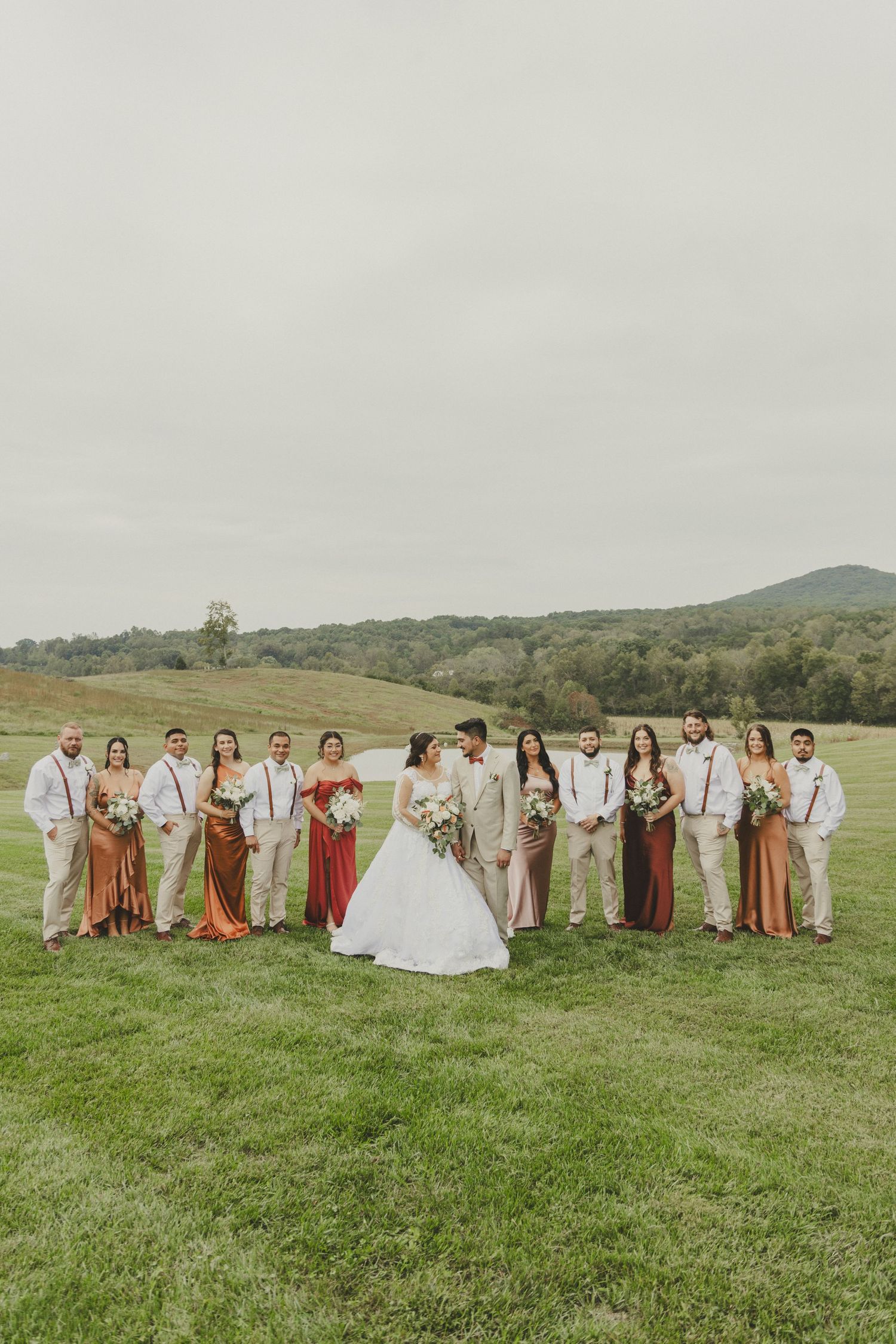 Wedding party in earth tones stands on grassy hillside with mountain backdrop on overcast autumn day.