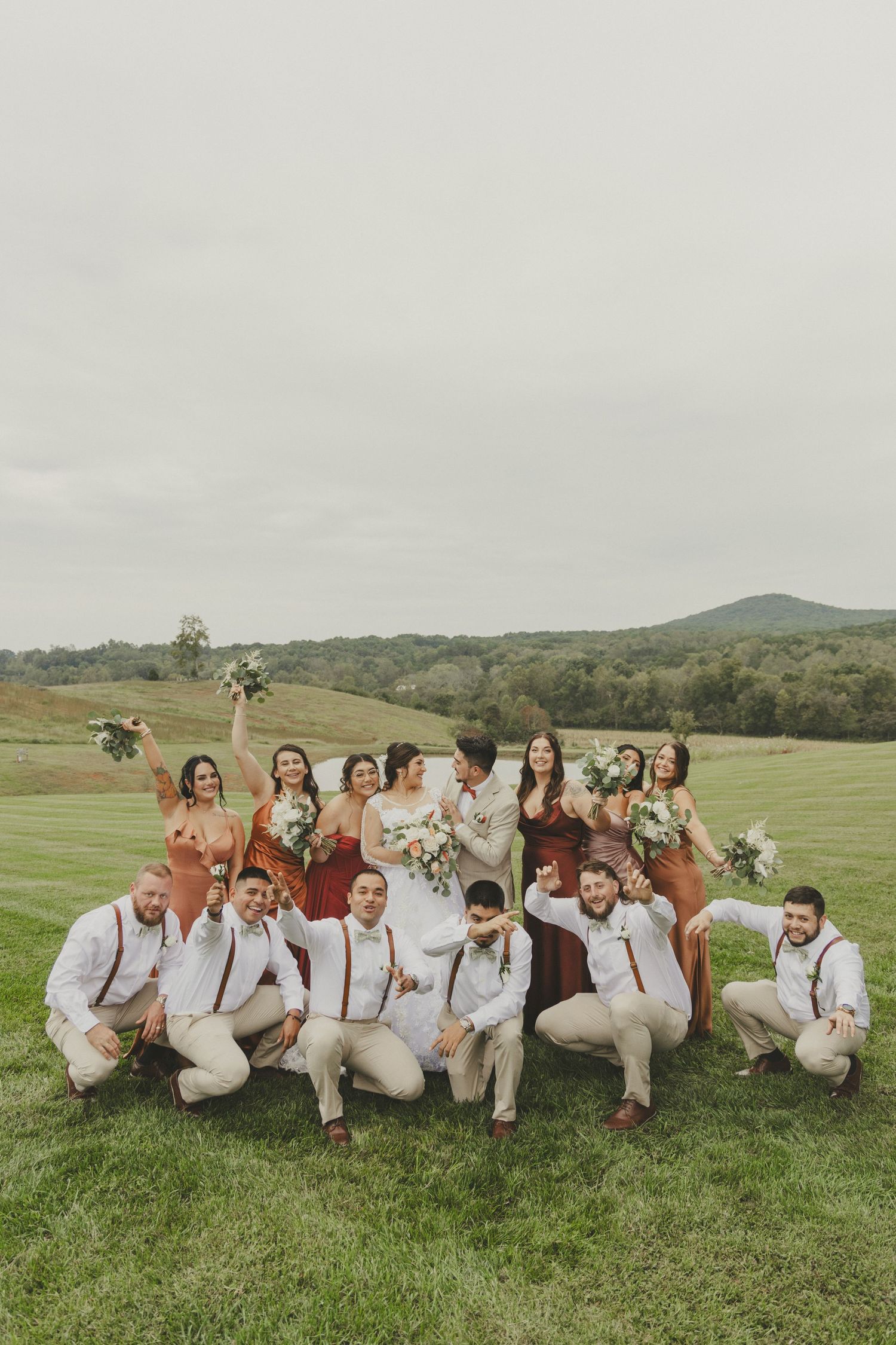 A wedding party poses playfully while sitting on grass in a scenic mountain setting.