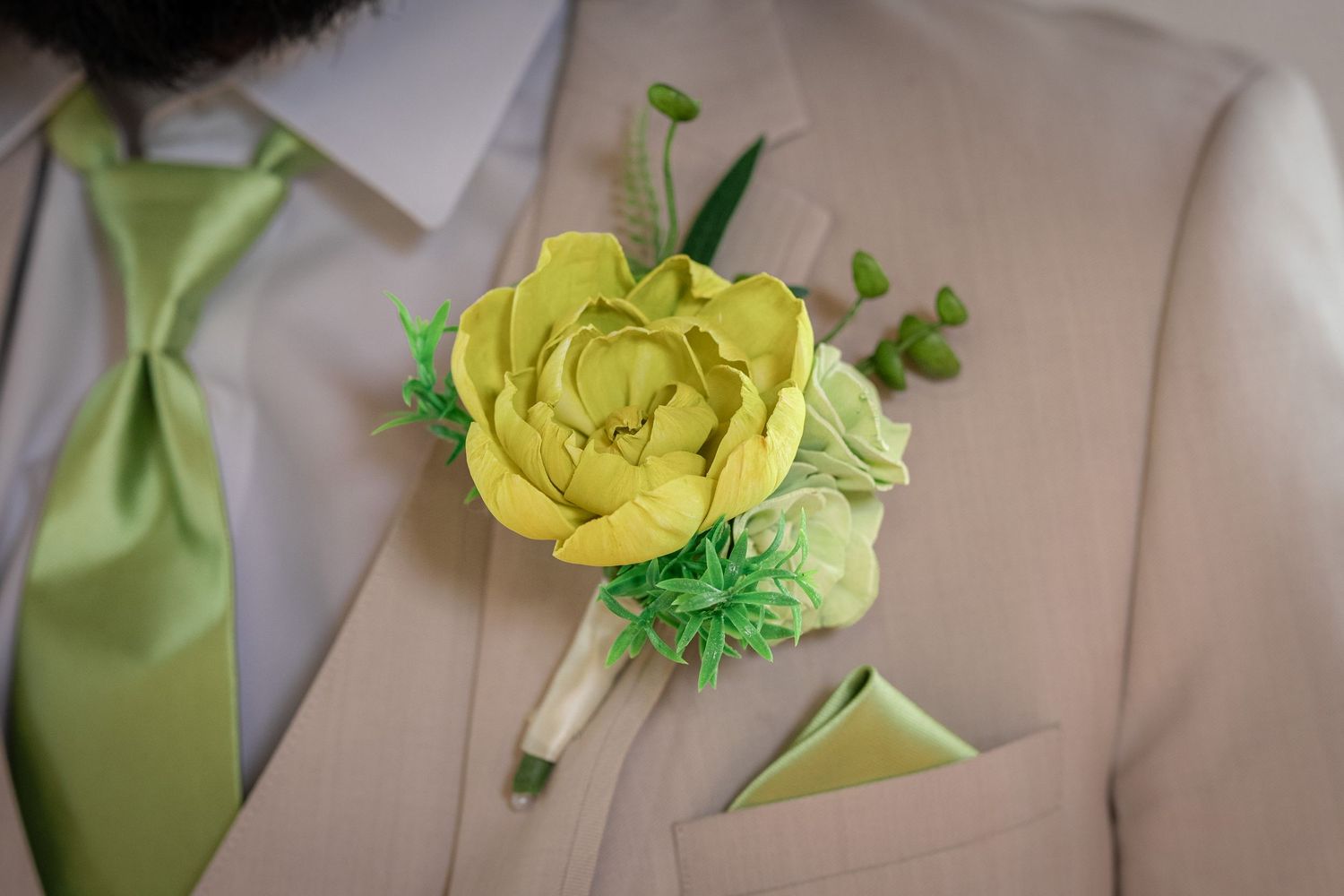 A vibrant yellow paper flower boutonniere with green accents pinned to a tan suit jacket beside a matching lime green tie.