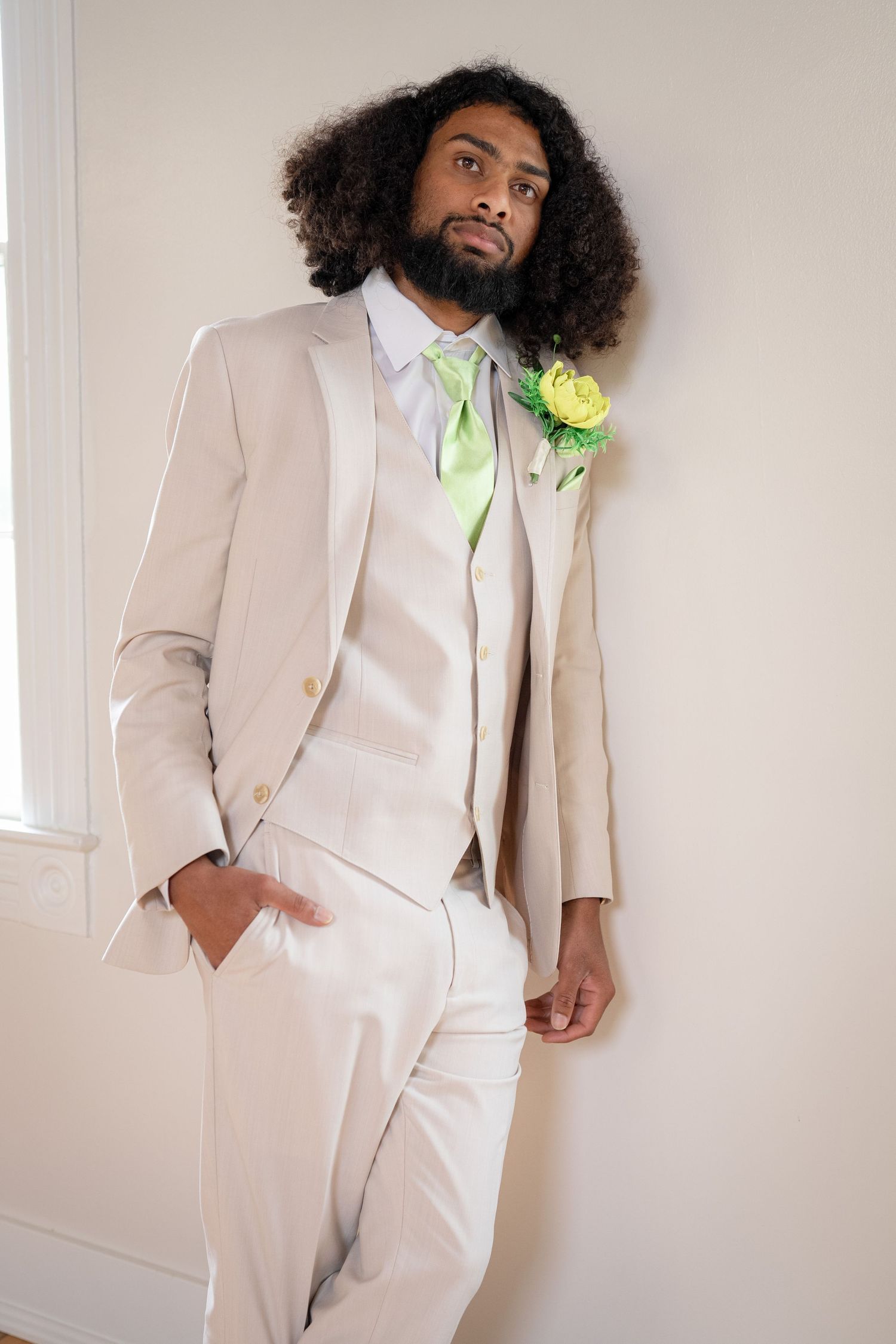 A person wearing a beige three-piece suit with a light green tie and boutonniere looks upward in an elegant pose.