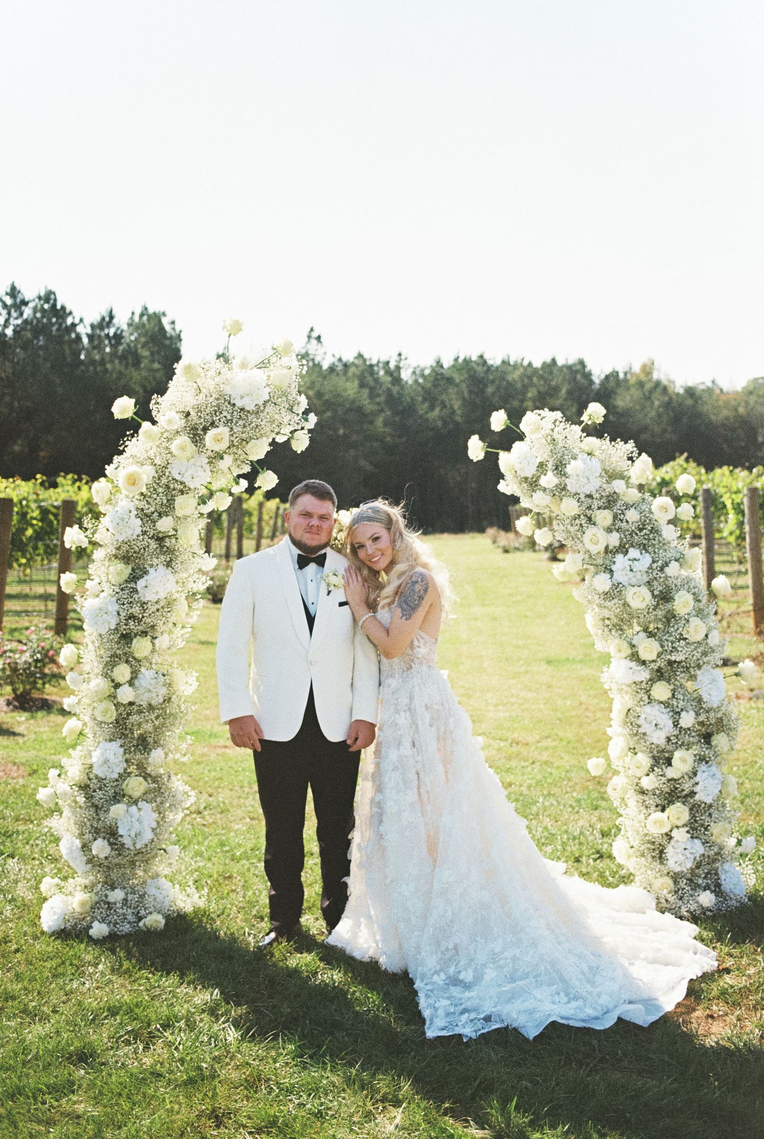A beautiful wedding ceremony setup with a floral arch made of white roses and greenery in an outdoor vineyard setting.