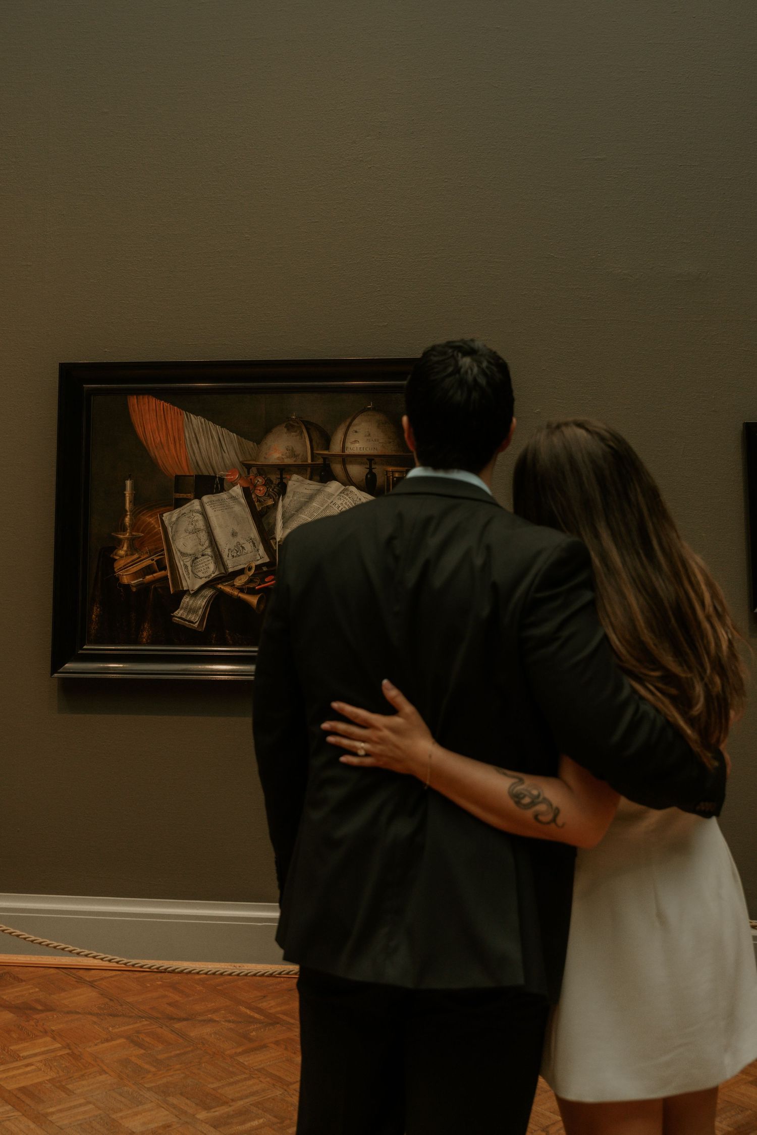 A couple observes artwork together in a gallery, standing close while viewing a framed piece on a dark wall.