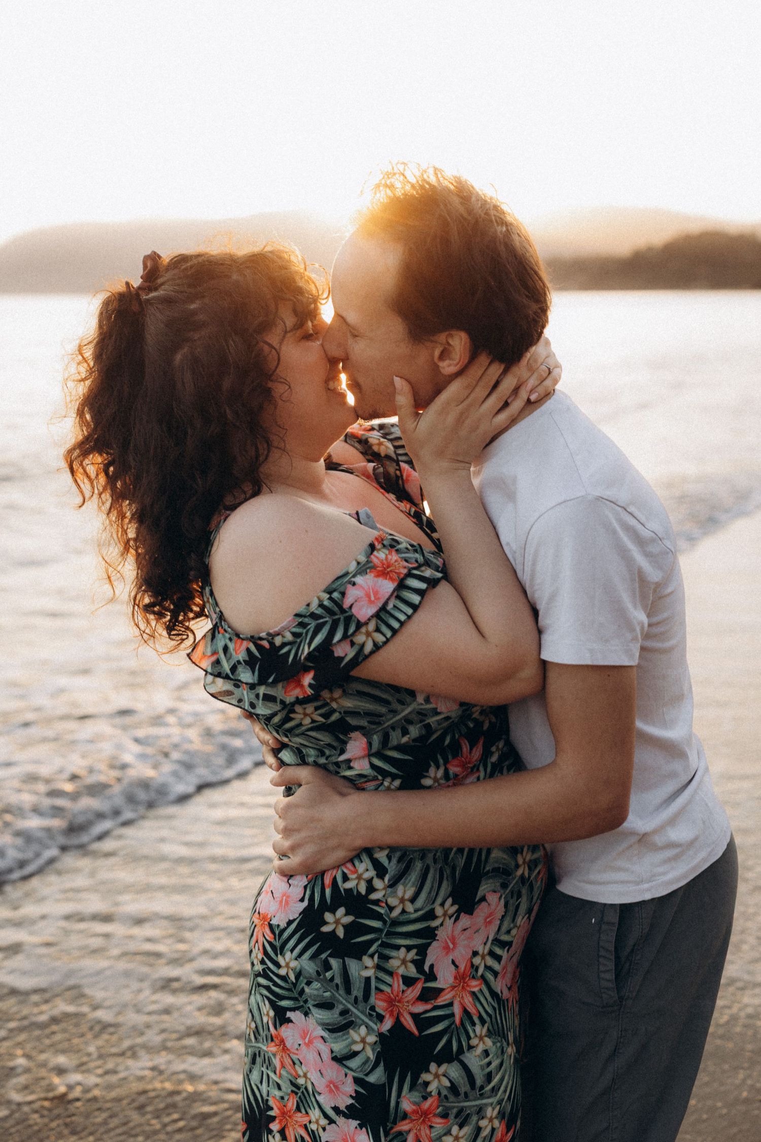 A couple shares a passionate kiss at sunset by the water, with warm light glowing behind them as waves sparkle nearby.