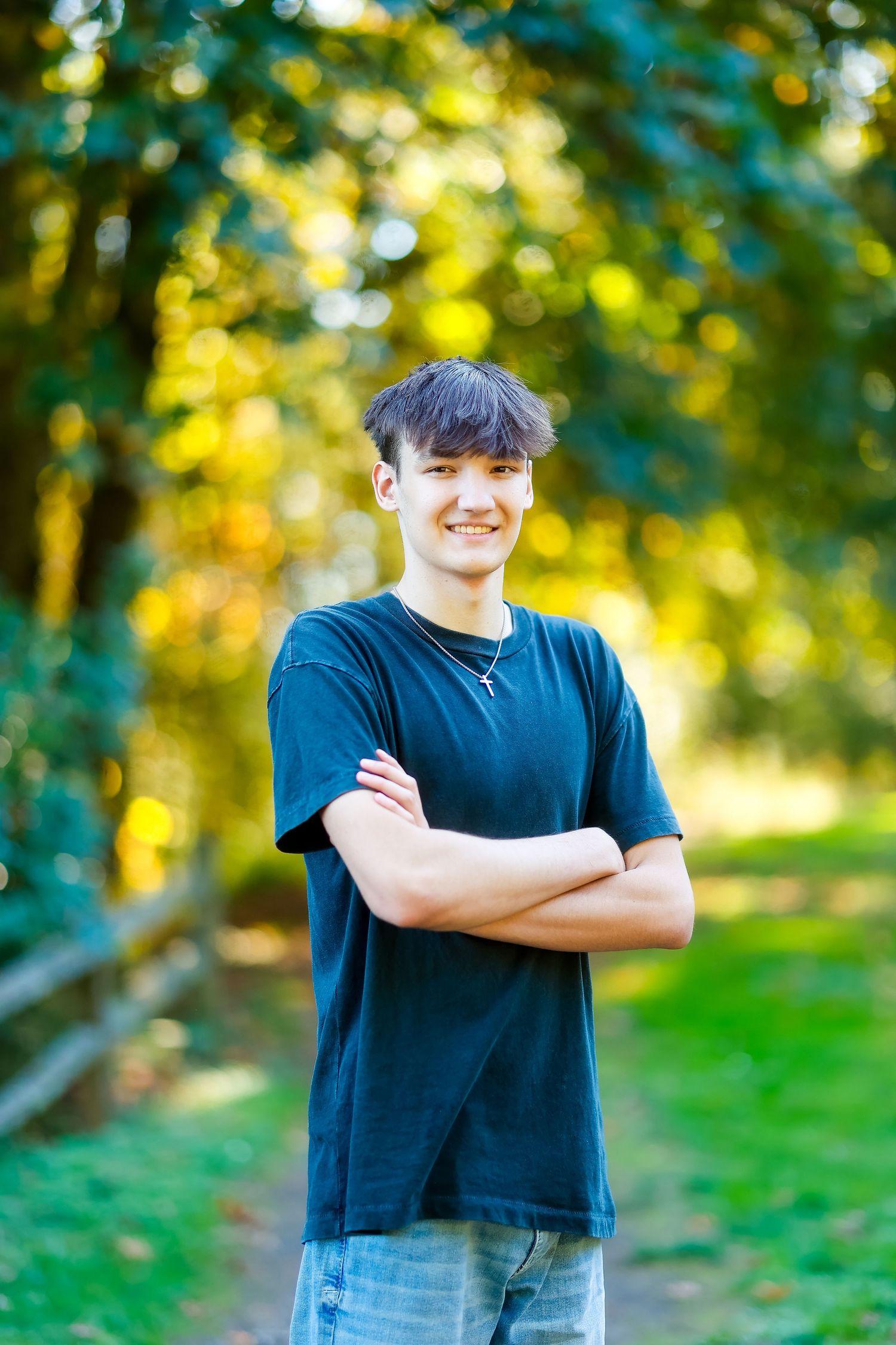 Person in navy t-shirt stands with arms crossed in sunny outdoor setting with blurred trees in background.
