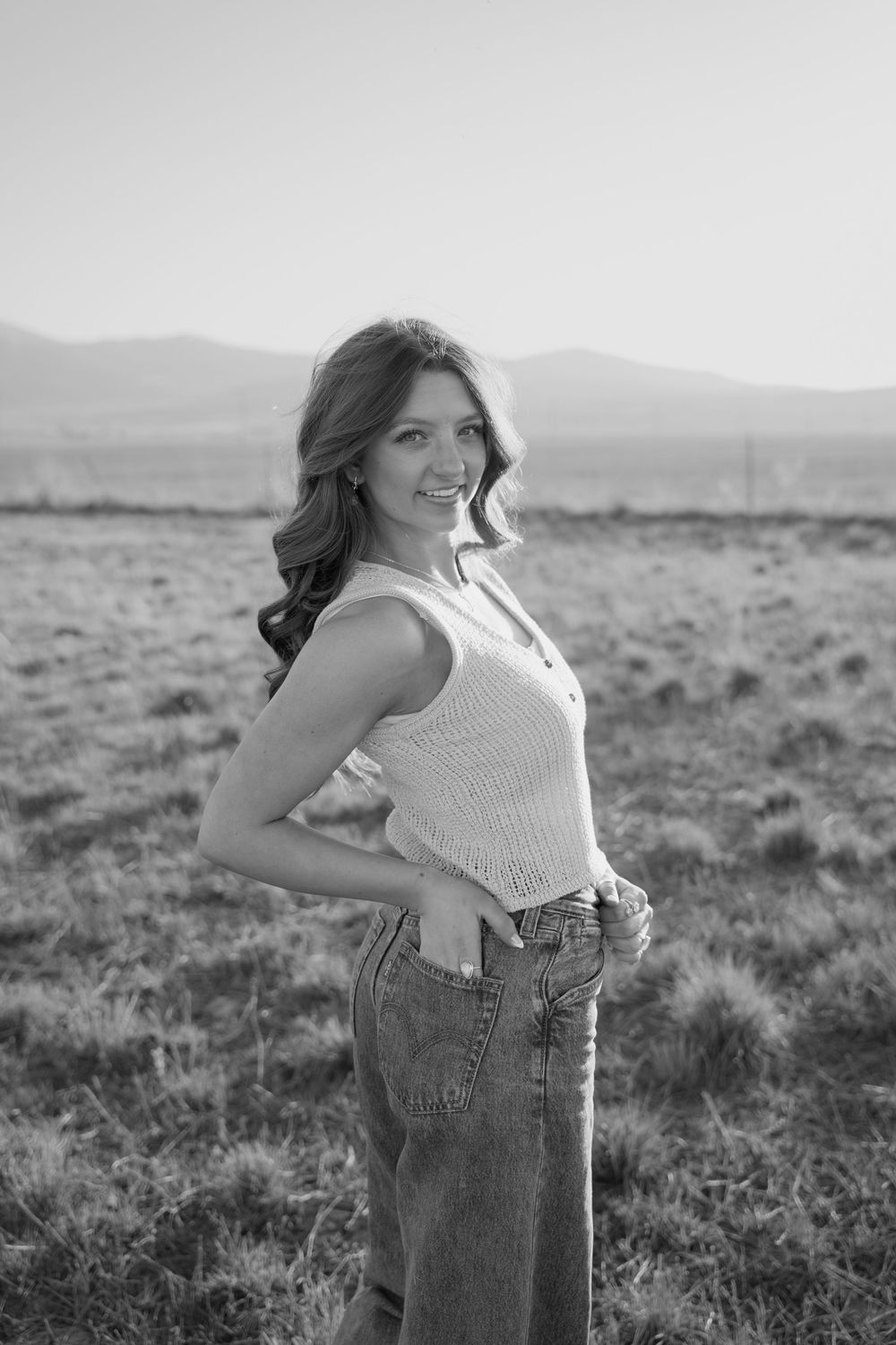 Black and white outdoor portrait of someone in a sleeveless top and jeans posing against a mountainous landscape.
