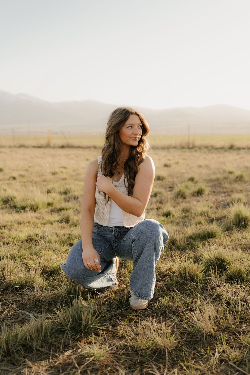 Person in casual white top and jeans poses in kneeling position against golden grassy field during sunset.