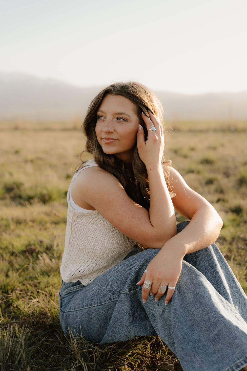 Casual portrait of someone wearing a cream tank top and jeans sitting on grass in a golden field at sunset.