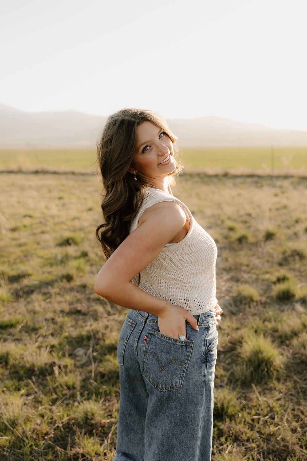 A person stands in a white sleeveless top and denim jeans against a scenic rural field during golden hour.