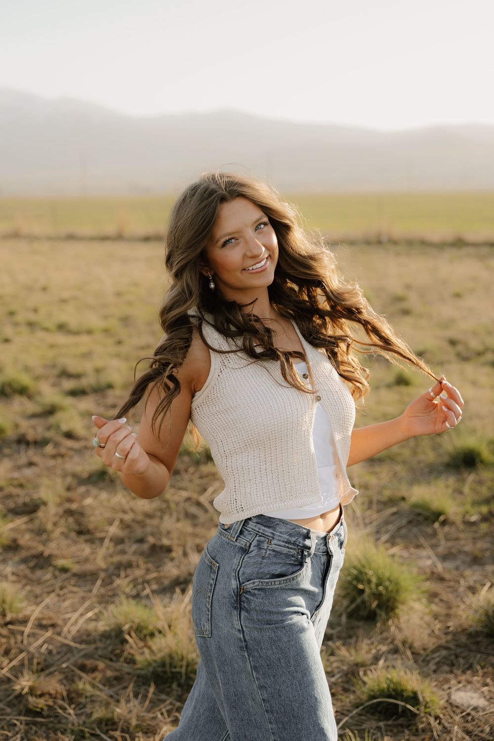 A person spins joyfully in a white top and jeans against a desert mountain landscape during golden hour.