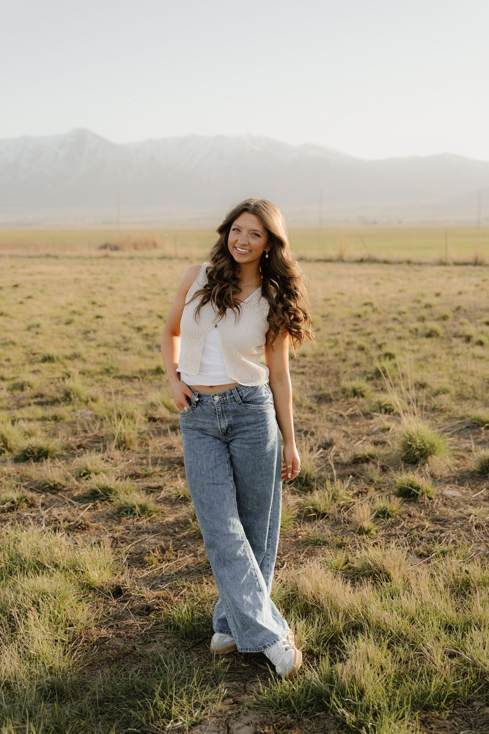 A casual outdoor portrait in a white crop top and wide-leg jeans against a mountainous landscape at golden hour.
