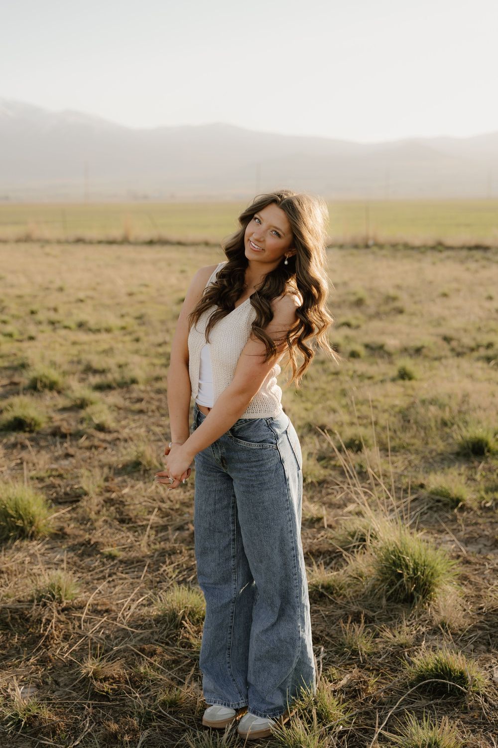 Someone wearing a white top and wide-leg jeans poses in a golden field at sunset with mountains in the background.