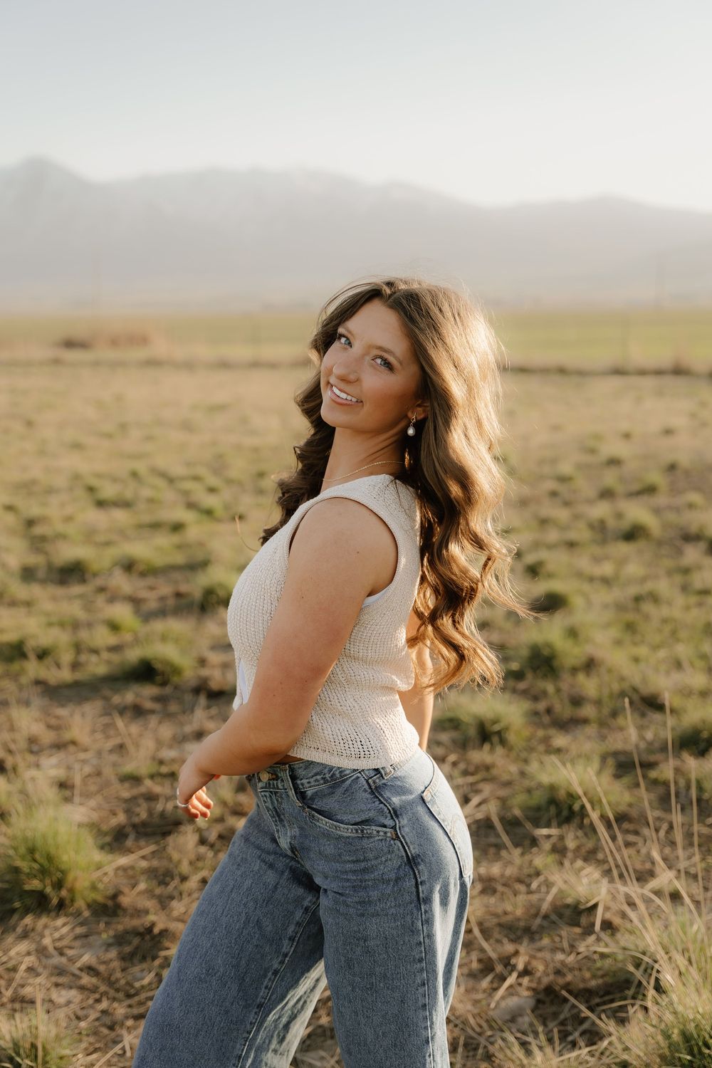 A person poses playfully in a field at sunset wearing denim overalls and a white top against a desert landscape.