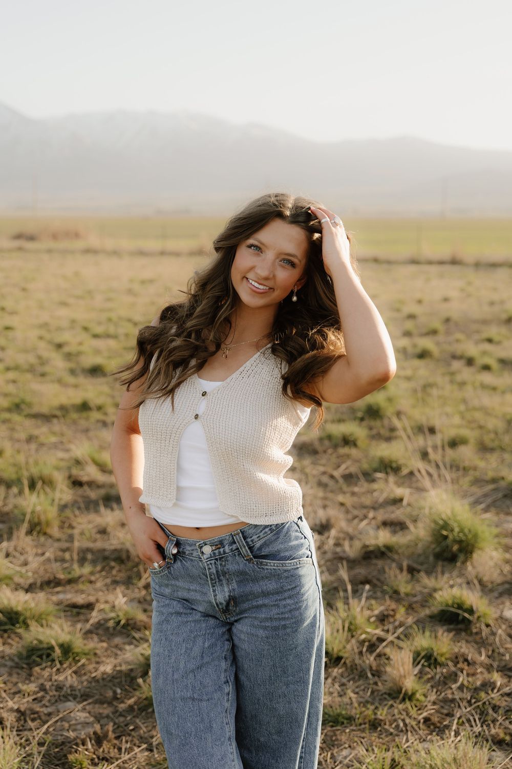 Casual portrait in an open field at sunset wearing white tank top and jeans against misty mountain backdrop.