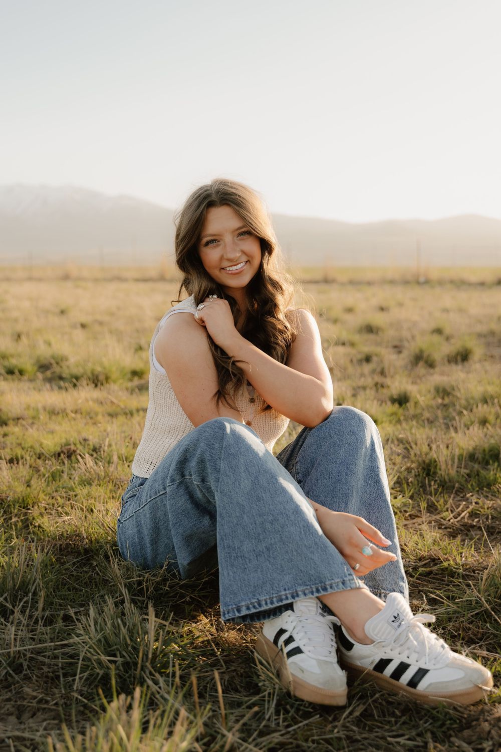 Someone in a white tank top and relaxed jeans sits in a golden field at sunset wearing Adidas sneakers.