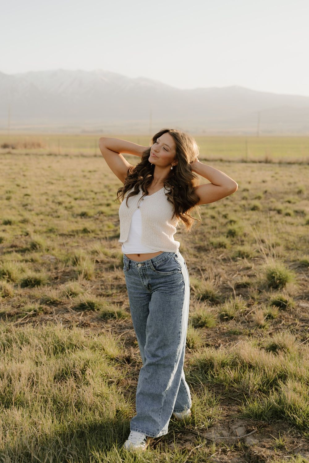 Figure in a white crop top and baggy jeans poses naturally in a grassy field at sunset with mountains in the distance.
