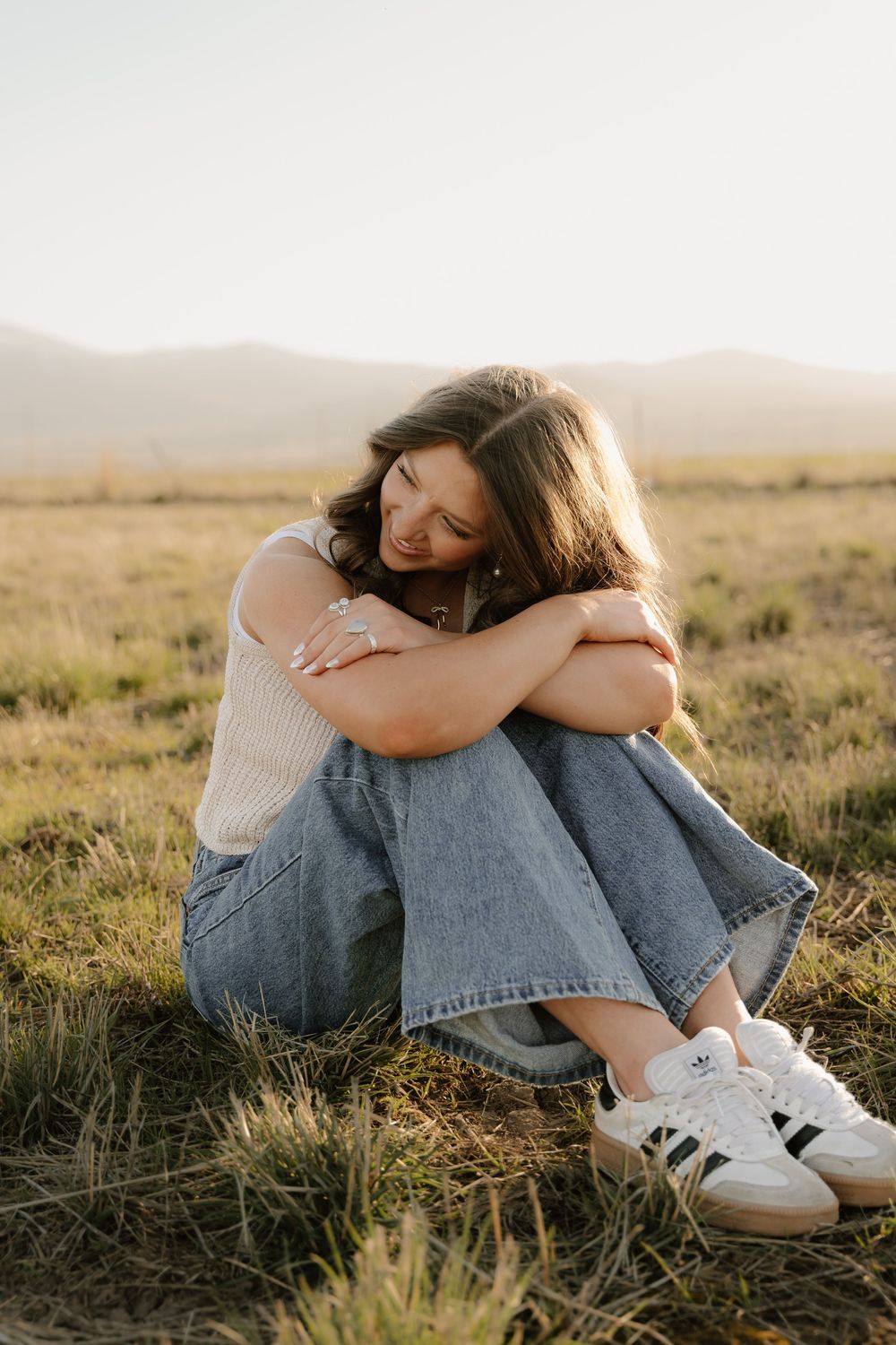 Young woman in white top and jeans sits in grassy field at sunset, hugging knees and smiling with eyes closed.