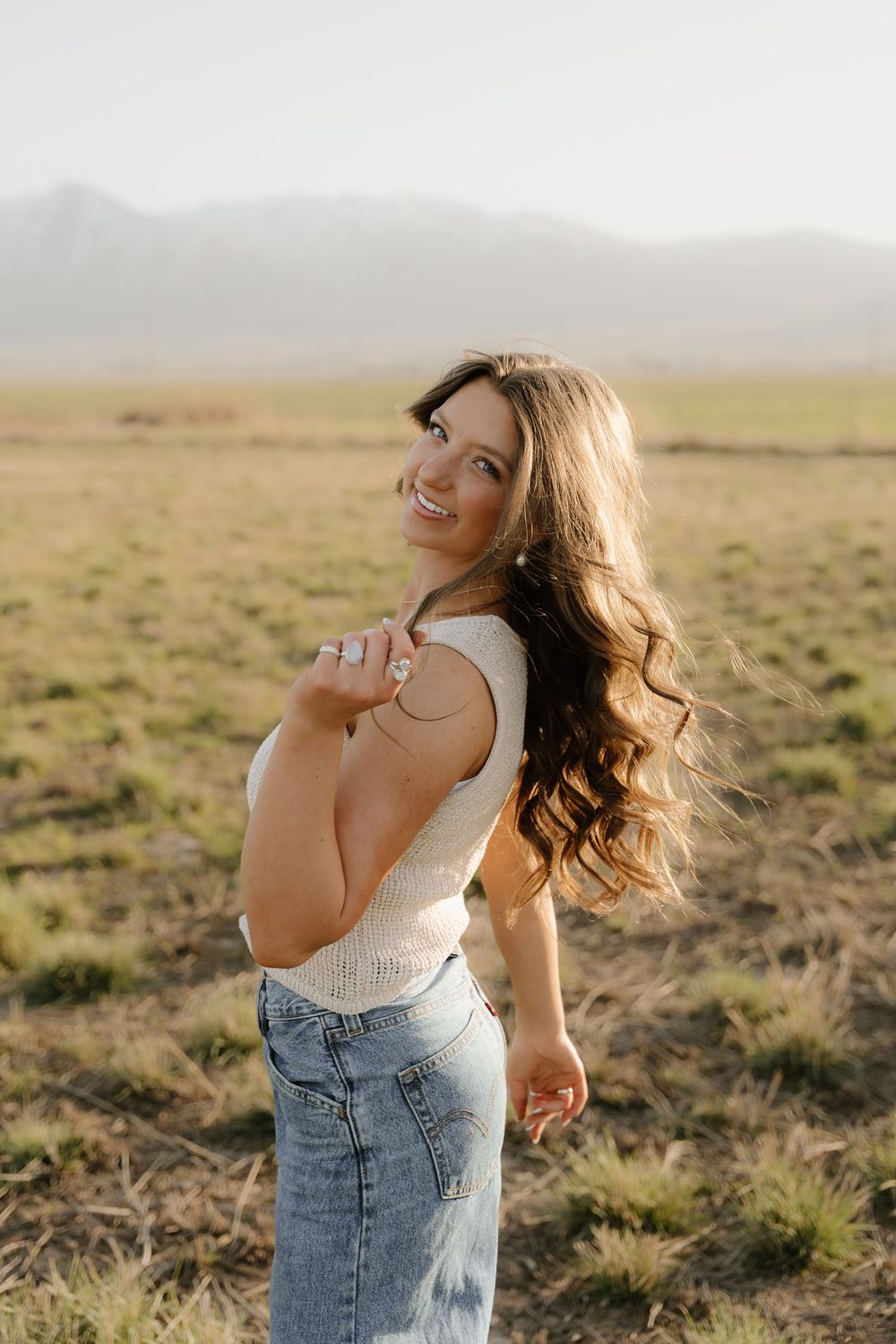 A person in a white top and jeans smiles while twirling with long wavy hair in a sunlit field with mountains.