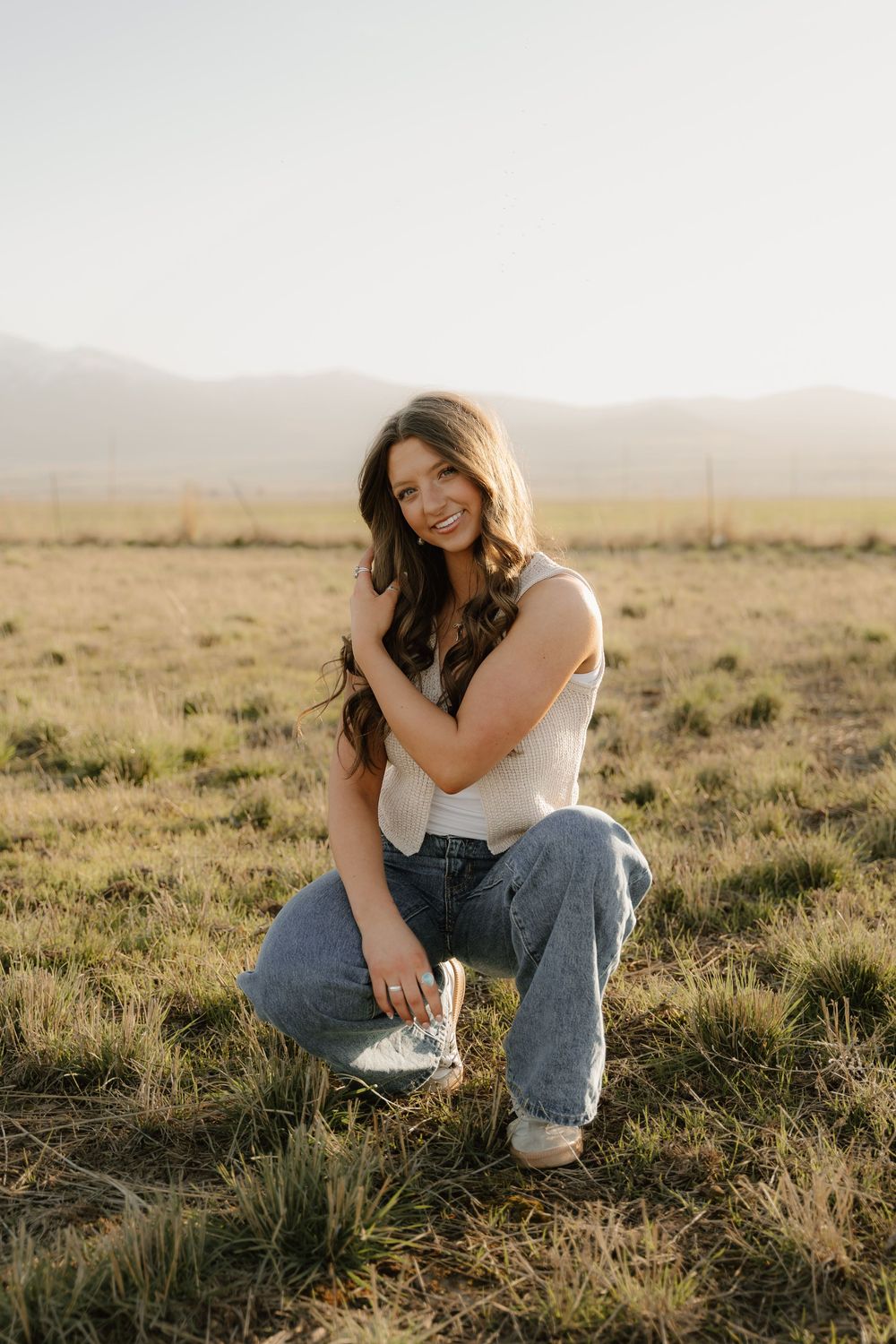 Person in white tank top and jeans crouches in golden field during sunset with mountains in background.