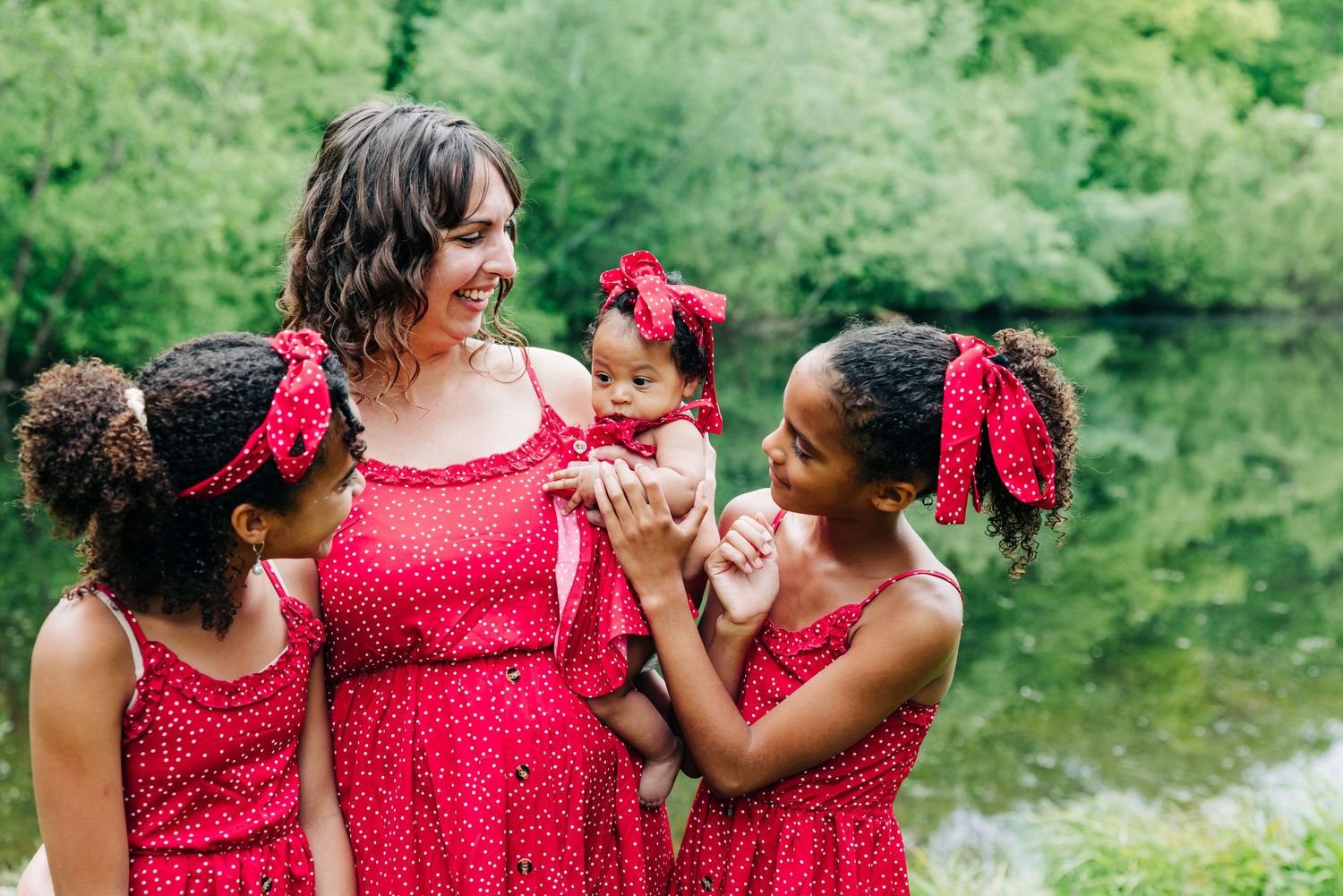 A family in matching red polka dot dresses with lace details share a joyful moment by a serene pond during a photo session at brooksvale