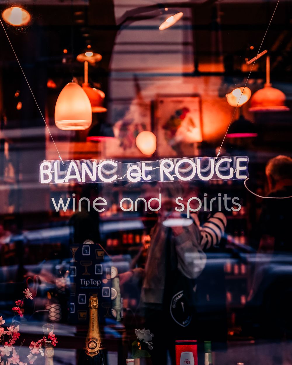 Neon sign glows in a wine shop window as warm lights and street reflections reveal layered moments of city life.