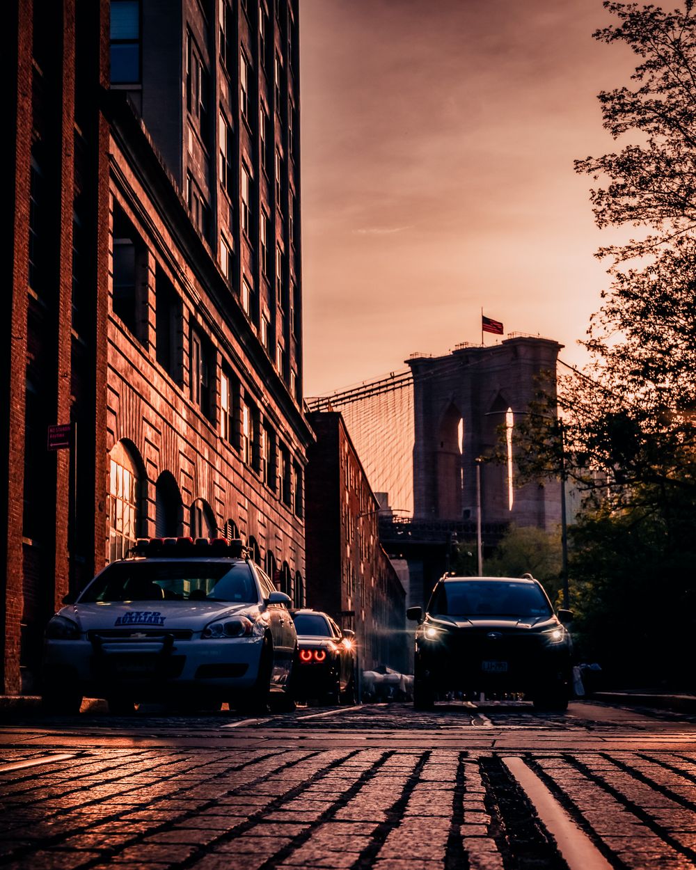 Cars idle on cobblestones at dusk beneath the Brooklyn Bridge, framed by golden light and old brick buildings.