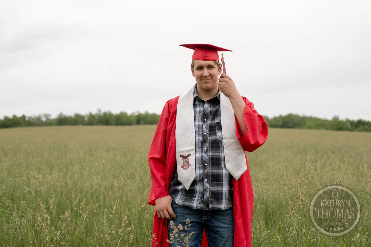 Graduate in red cap and gown poses in an open field on graduation day while wearing plaid shirt and jeans.