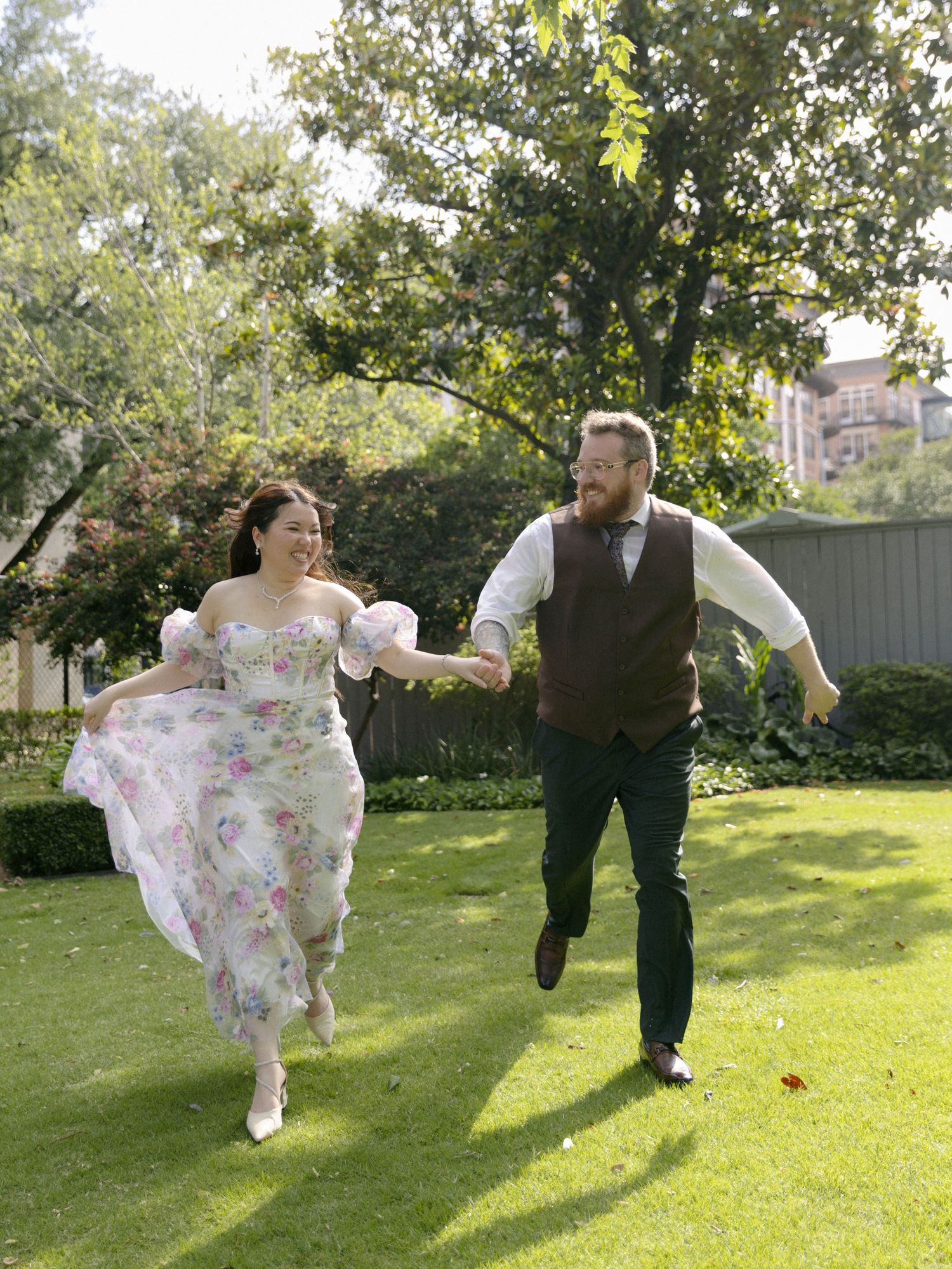 Two people in wedding attire run joyfully together on a sunny lawn surrounded by trees and greenery.