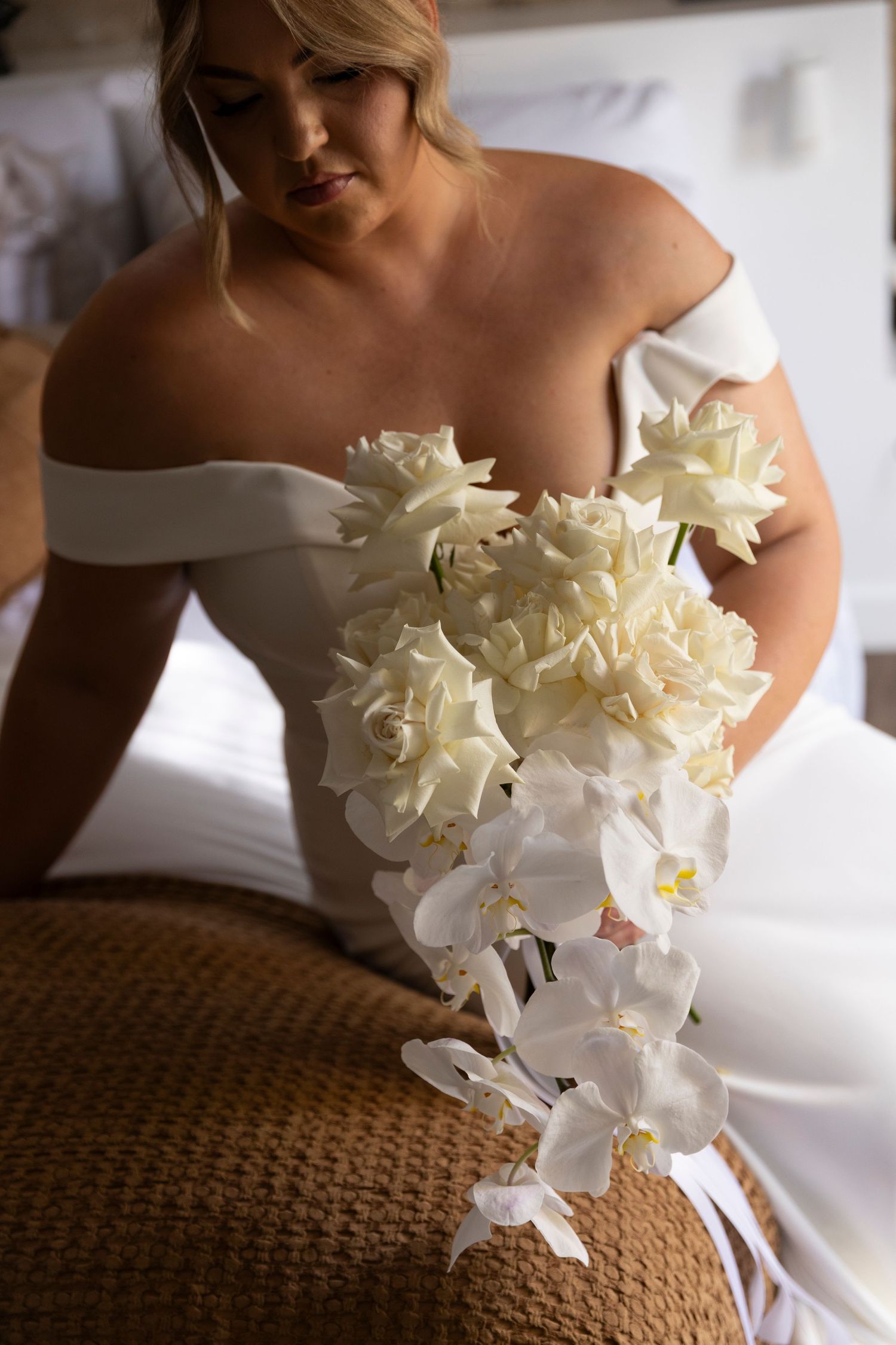 Bride in off-shoulder white dress holds elegant bouquet of white orchids and roses.