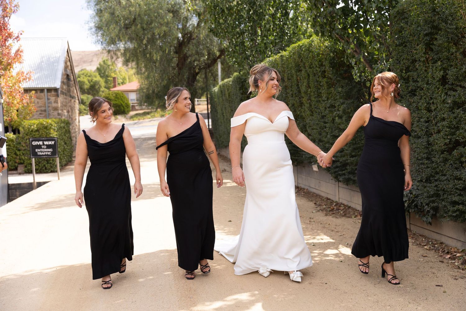 A wedding party in formal attire walks together holding hands along a path outdoors on a sunny day.