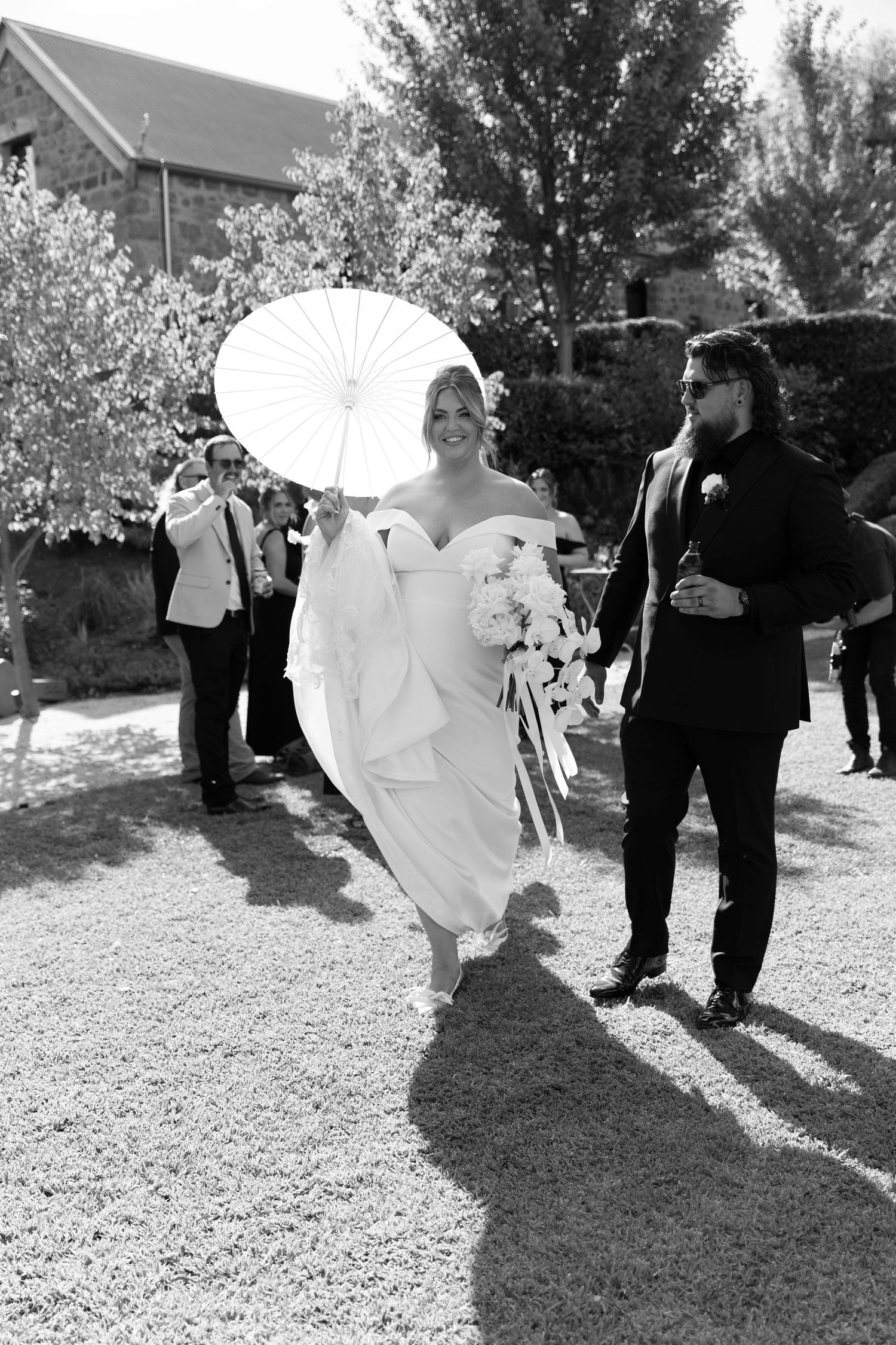 A bride holds a white parasol while walking with her partner during an outdoor wedding.