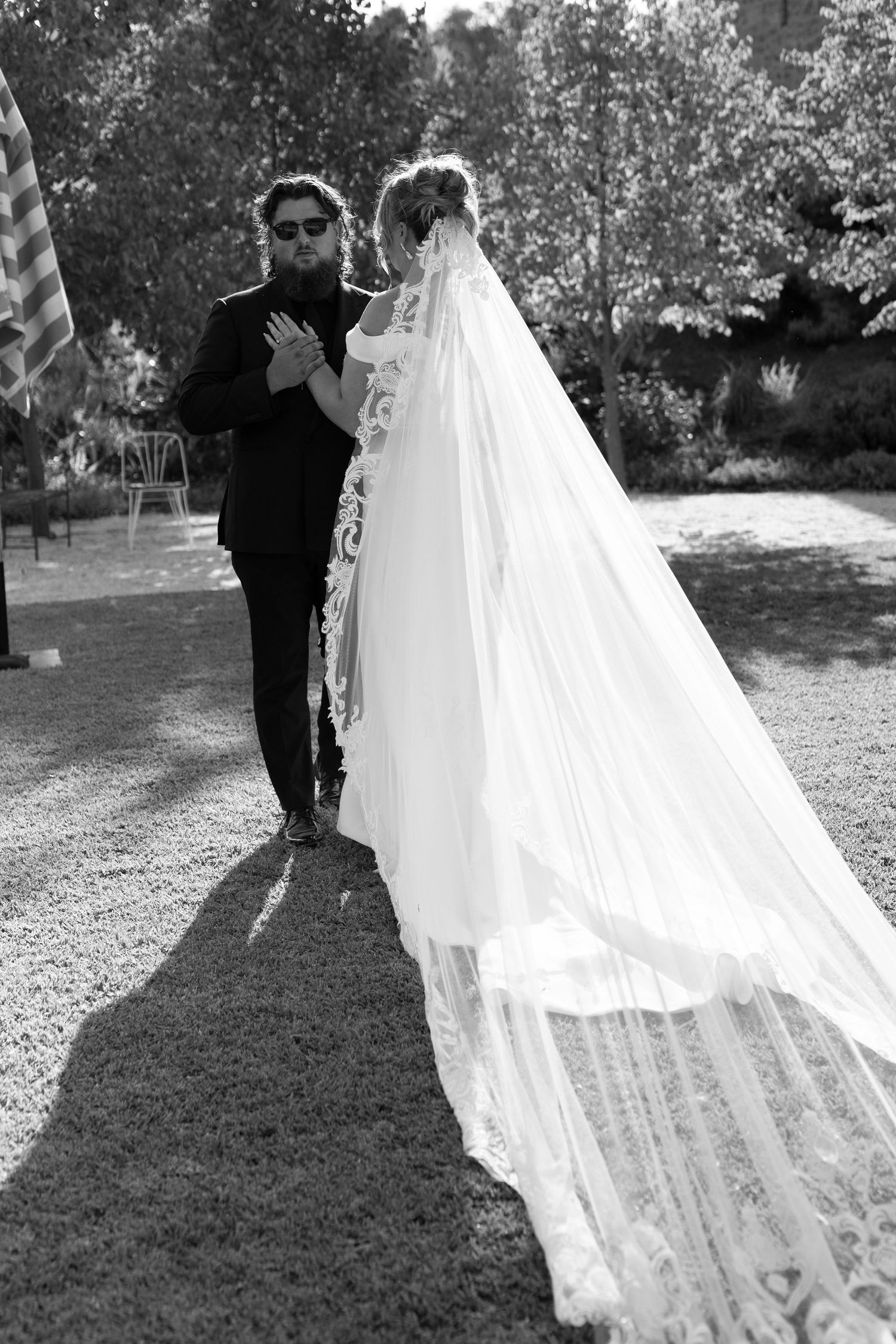 Couple shares romantic moment on garden path with flowing veil in black and white photo.