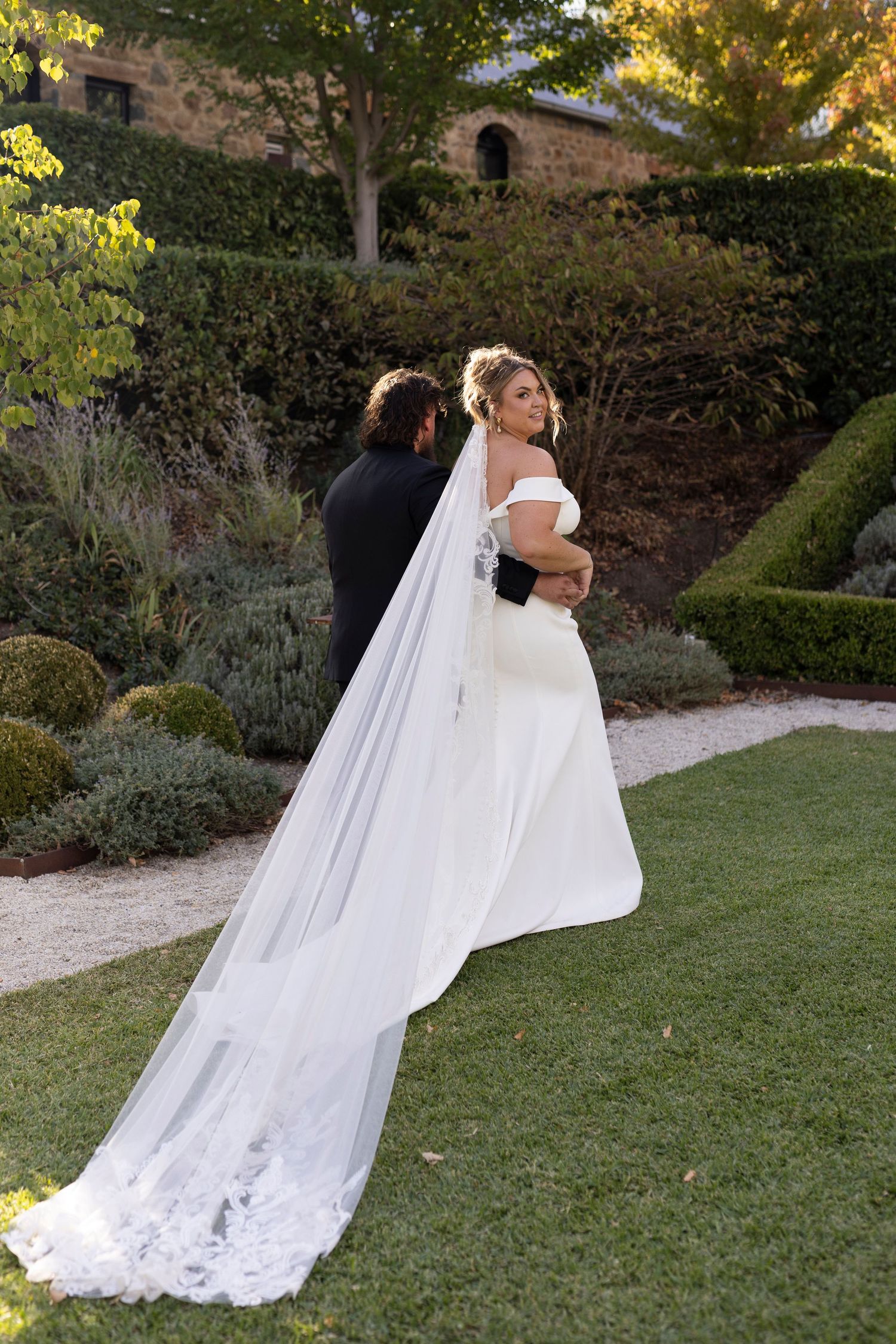 Couple embraces in garden setting with bride's long lace veil trailing behind.