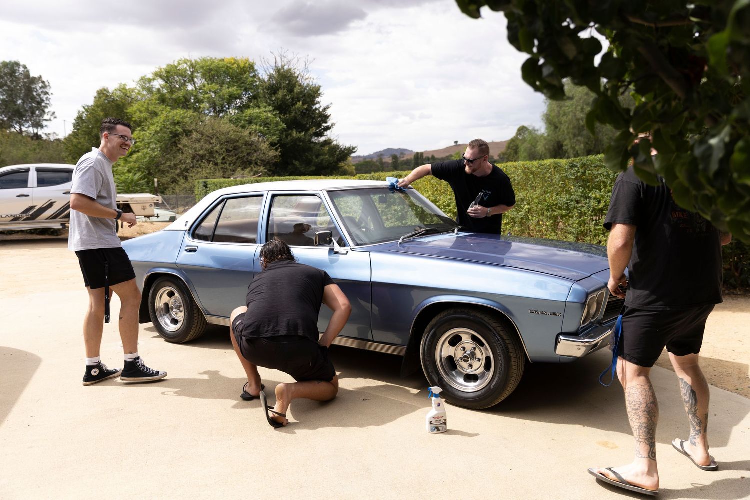 A group of people inspect a classic blue Opel car parked outdoors during a sunny day.