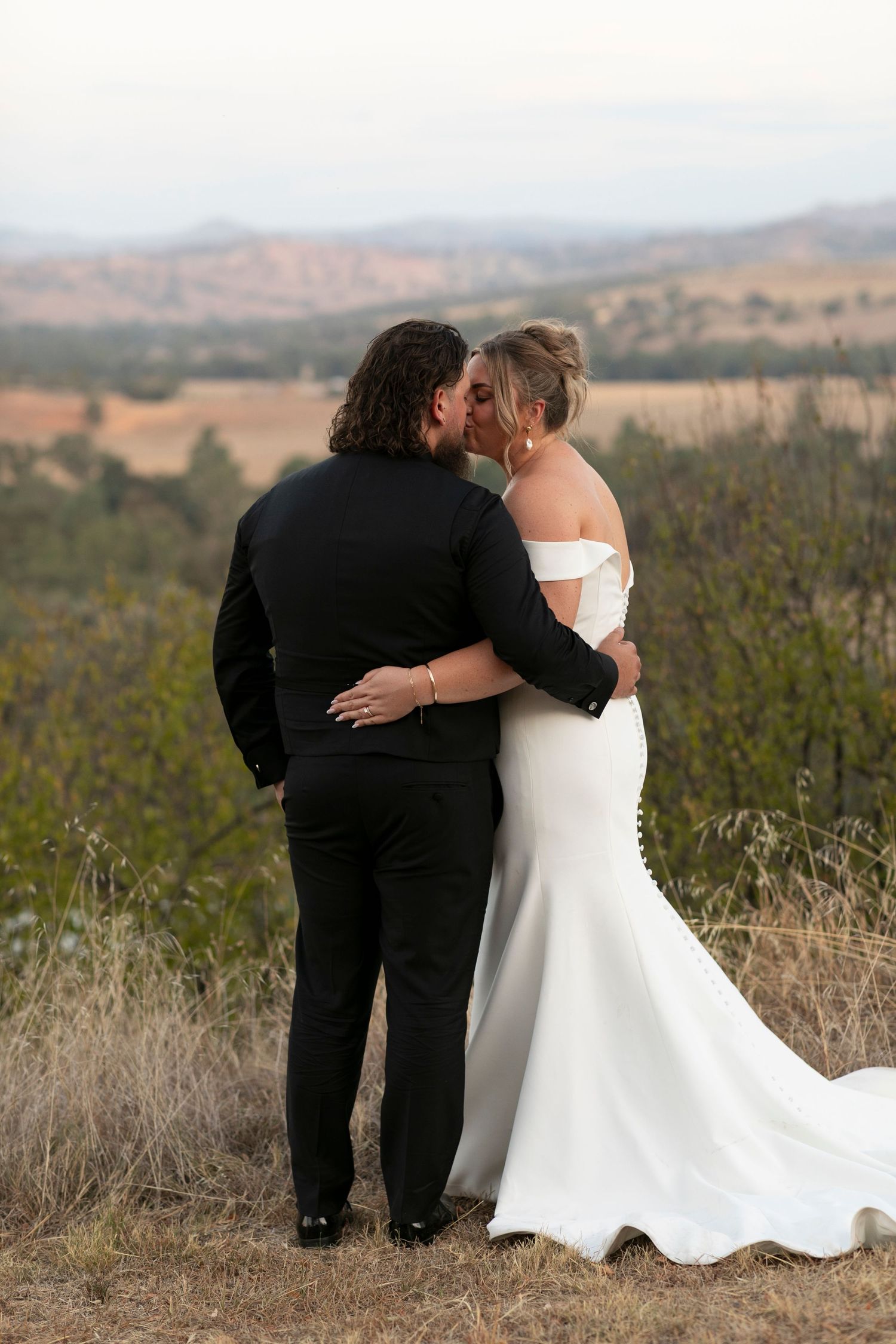 A couple in wedding attire embraces while overlooking a scenic mountain landscape at sunset.