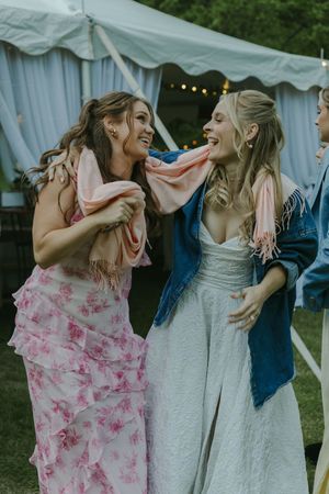 Two friends laugh joyfully at a wedding reception under a white tent, one in a pink floral dress and one in a white dress.