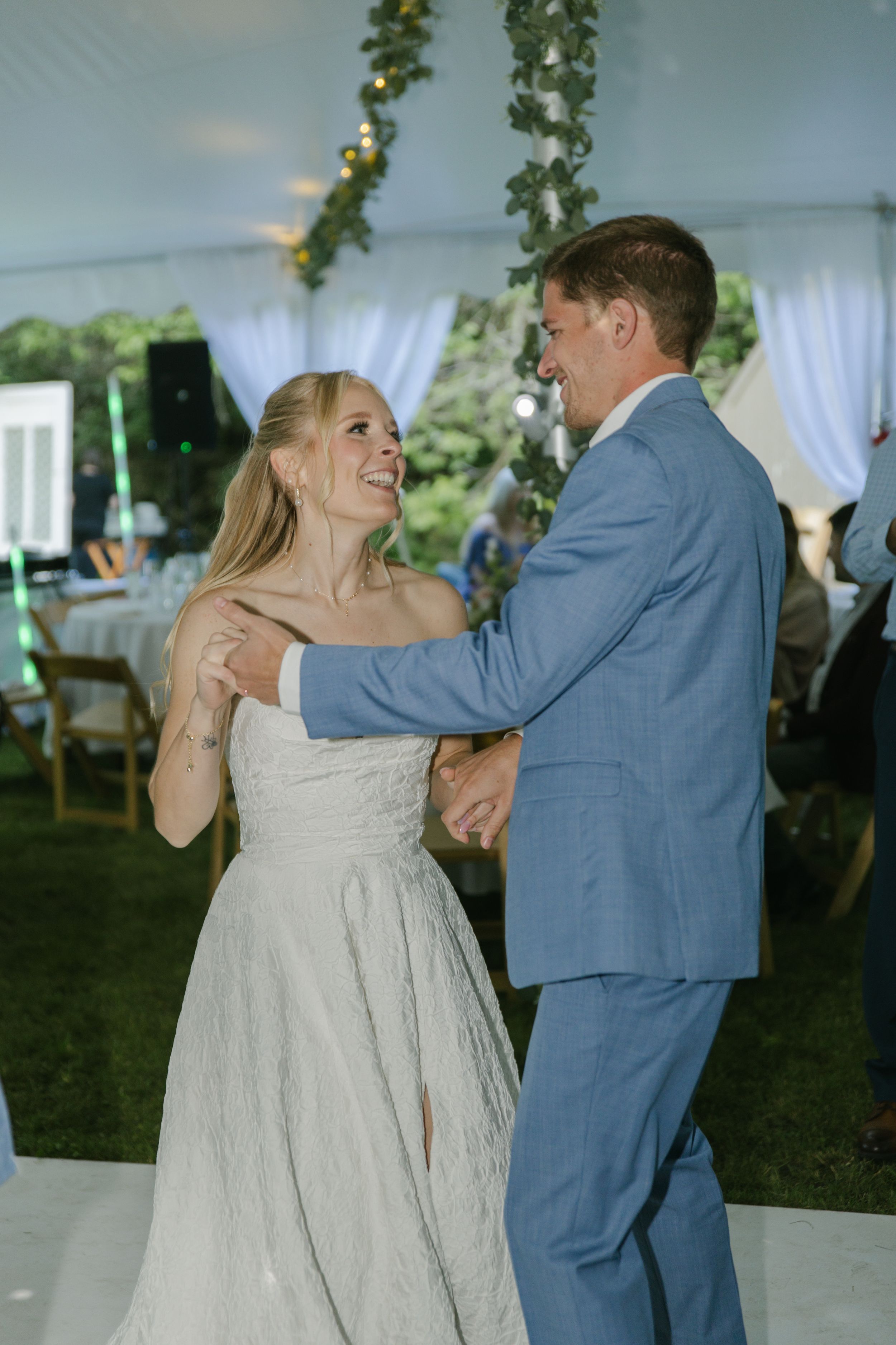 A couple shares their first dance under a tent decorated with green garlands and soft lighting.