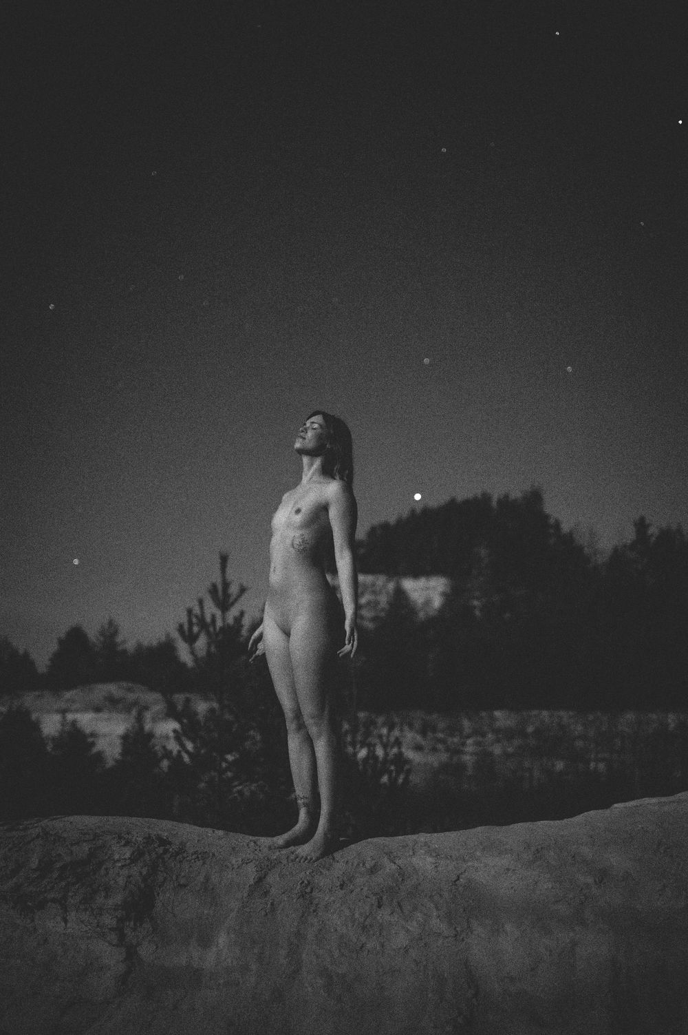 Moody grayscale photograph capturing figure standing on rocky outcrop under starlit sky.
