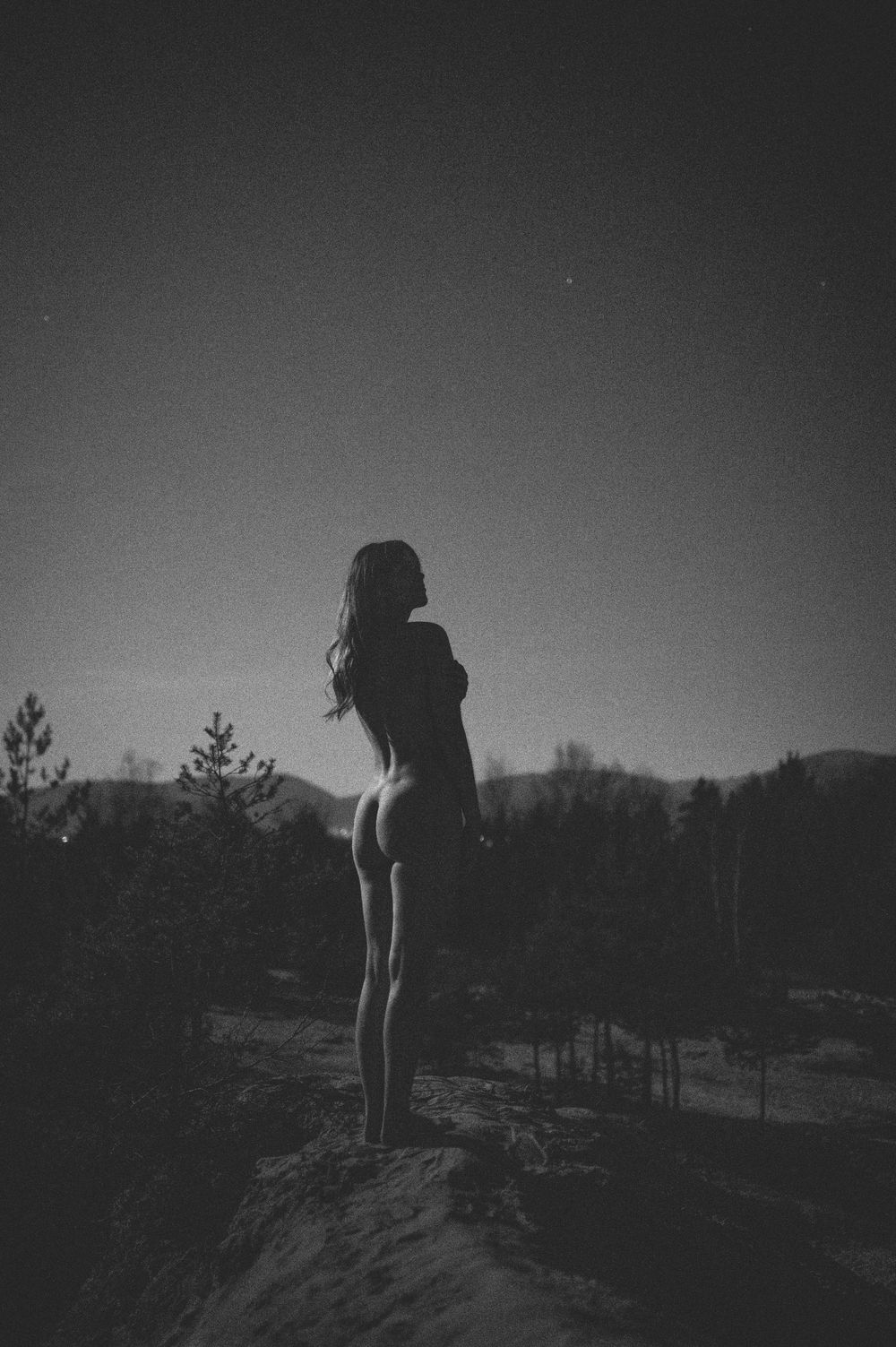Dramatic noir photograph showing silhouetted figure atop cliff edge with pine forest backdrop.